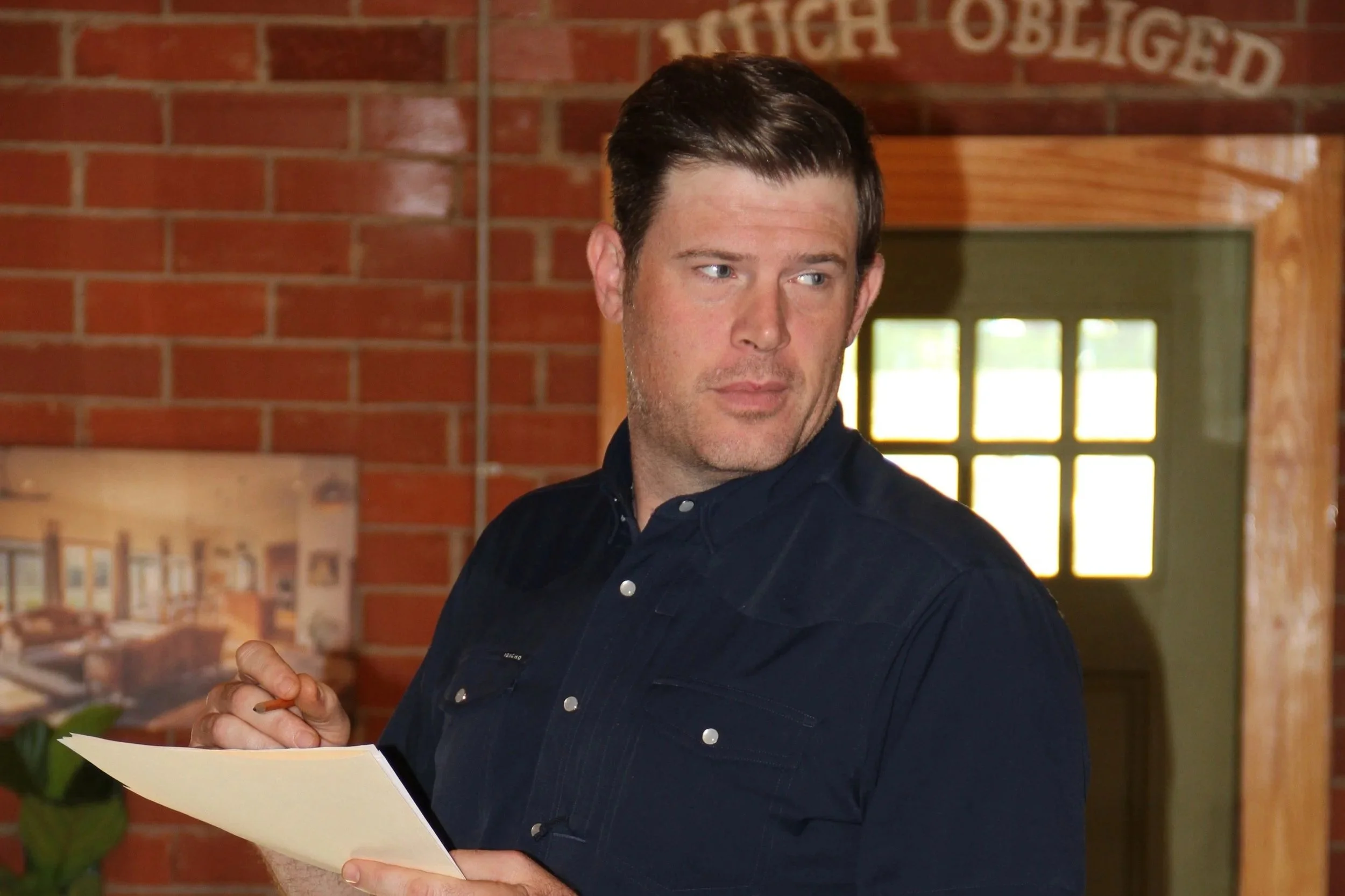 A man with dark hair and light skin, wearing a black button-up shirt, is holding a sheet of paper and looking to the side. The background features a red brick wall and a window with sunlight coming through.