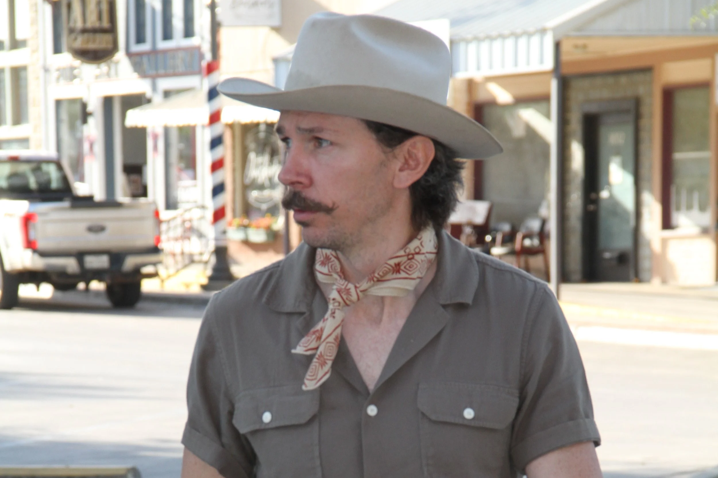 A man wearing a white cowboy hat, a patterned scarf, and a gray shirt walking outdoors in a small town or shopping district during daytime.