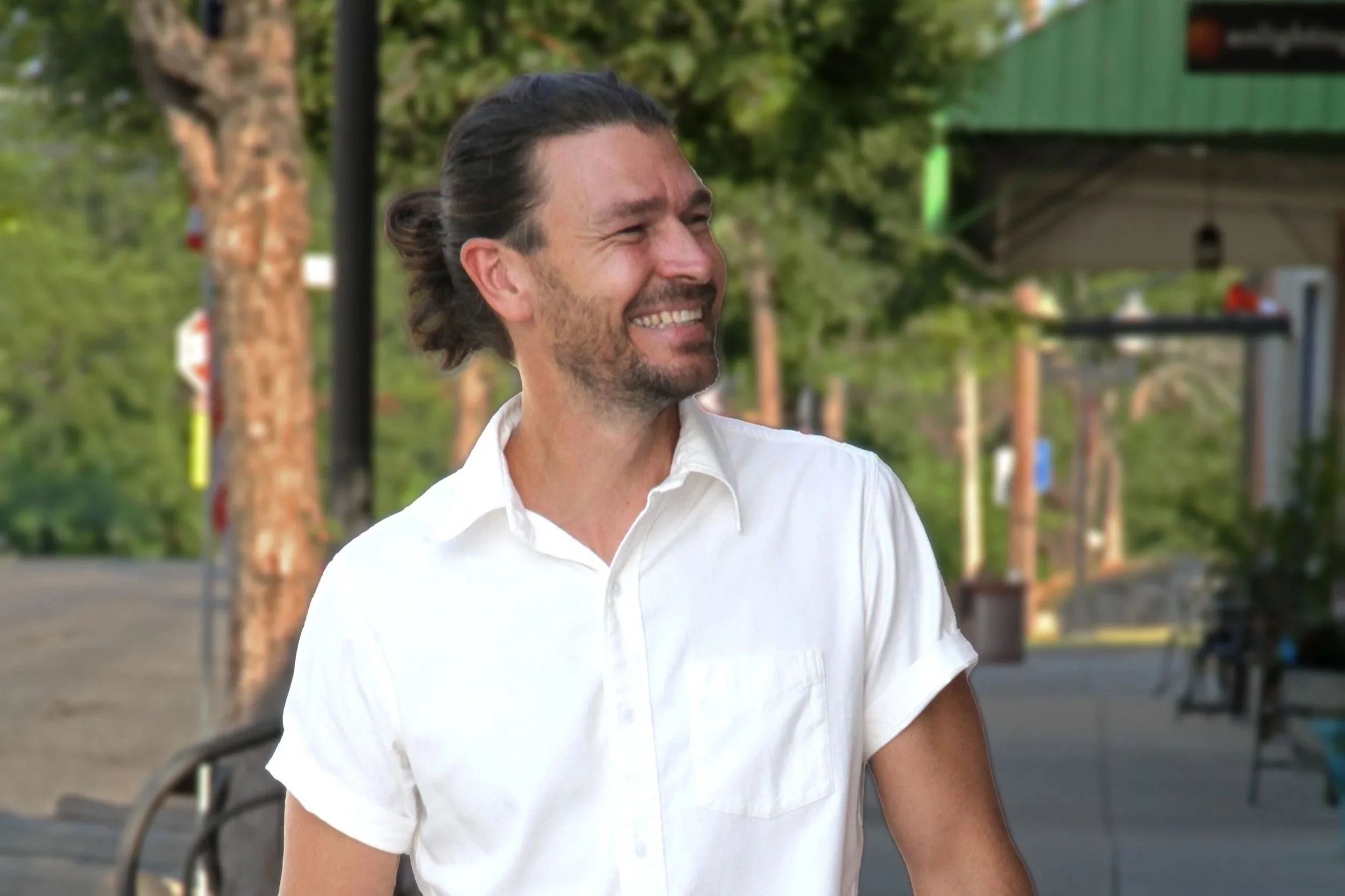 A man with dark hair tied back in a ponytail and a beard, smiling and wearing a white shirt, outdoors on a sunny day with green trees and a building in the background.
