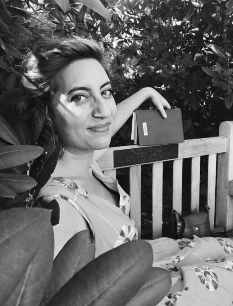 A portrait image of a woman in a dress sitting on a bench with leaves nearby