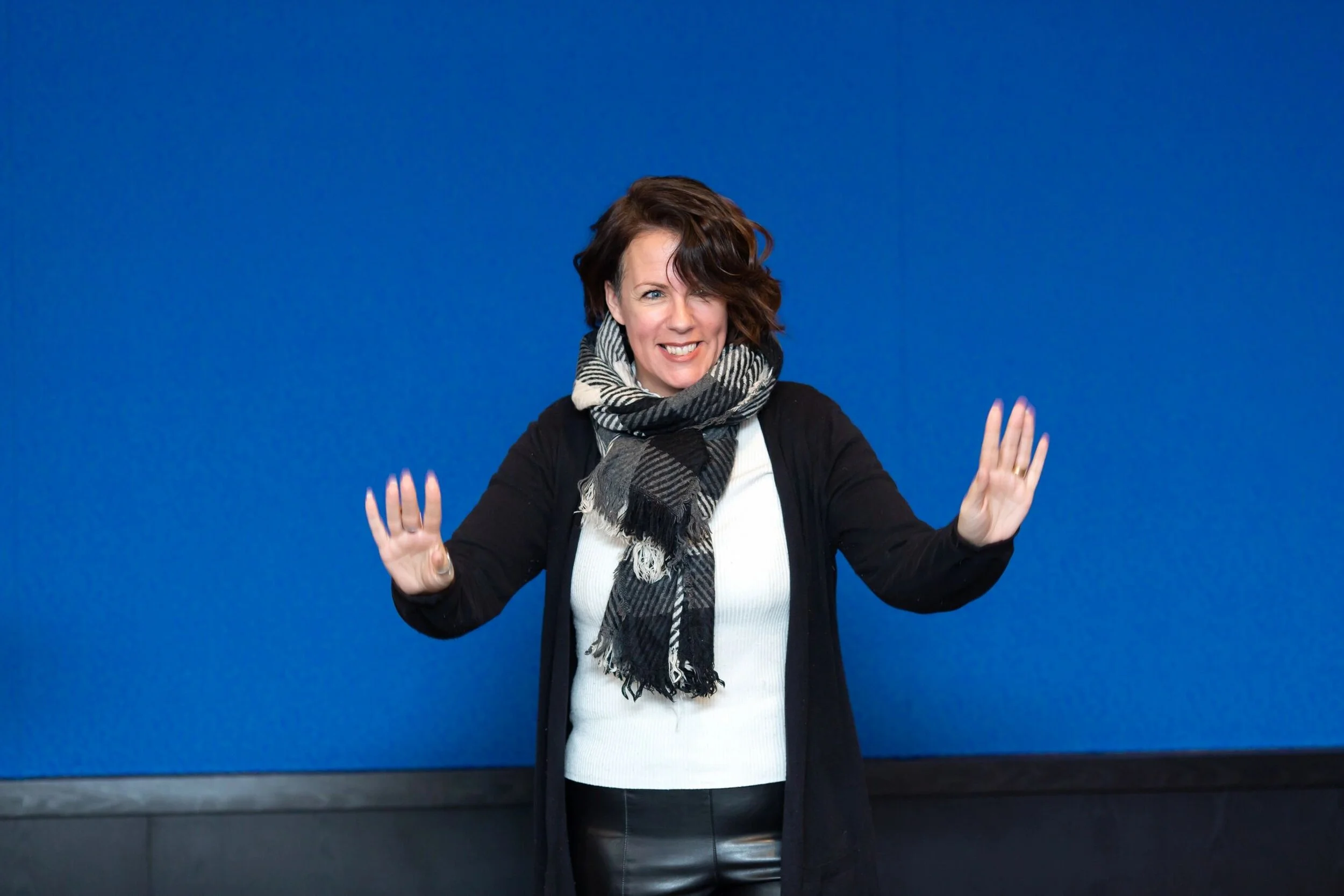 Lisa is pictured wearing a black sweater, white shirt, and black and white scarf. Her hands are up while she is speaking and she is smiling.  She has dark, shoulder length hair.