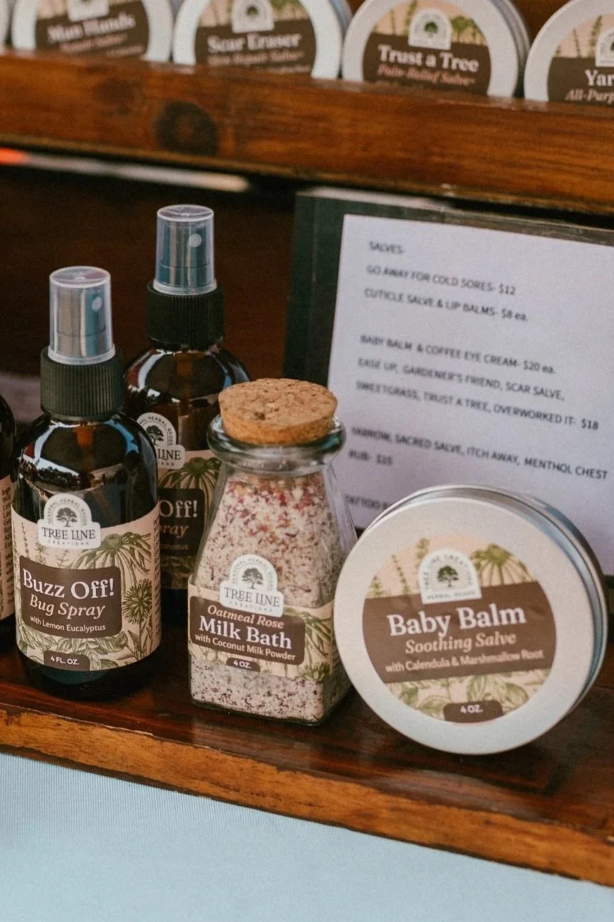 A small tin container labeled 'Tree Line Creations Lip Pomade Spring' on a wooden surface. Next to it is a glass jar with a cork lid containing a pinkish substance, possibly a bath or beauty product. Other jars and containers are blurred in the background, all placed on a crocheted lace doily.