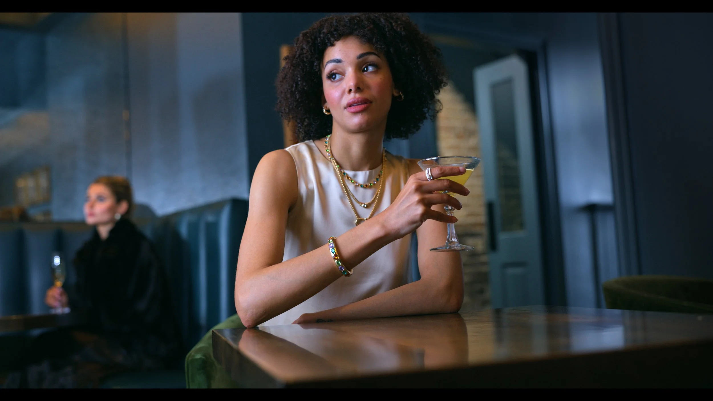 A woman with curly hair and jewelry, wearing a satin top, holding a martini glass and looking to the side in a dimly lit modern bar or restaurant. In the background, another woman is seated, slightly blurred, with dark hair and clothing.