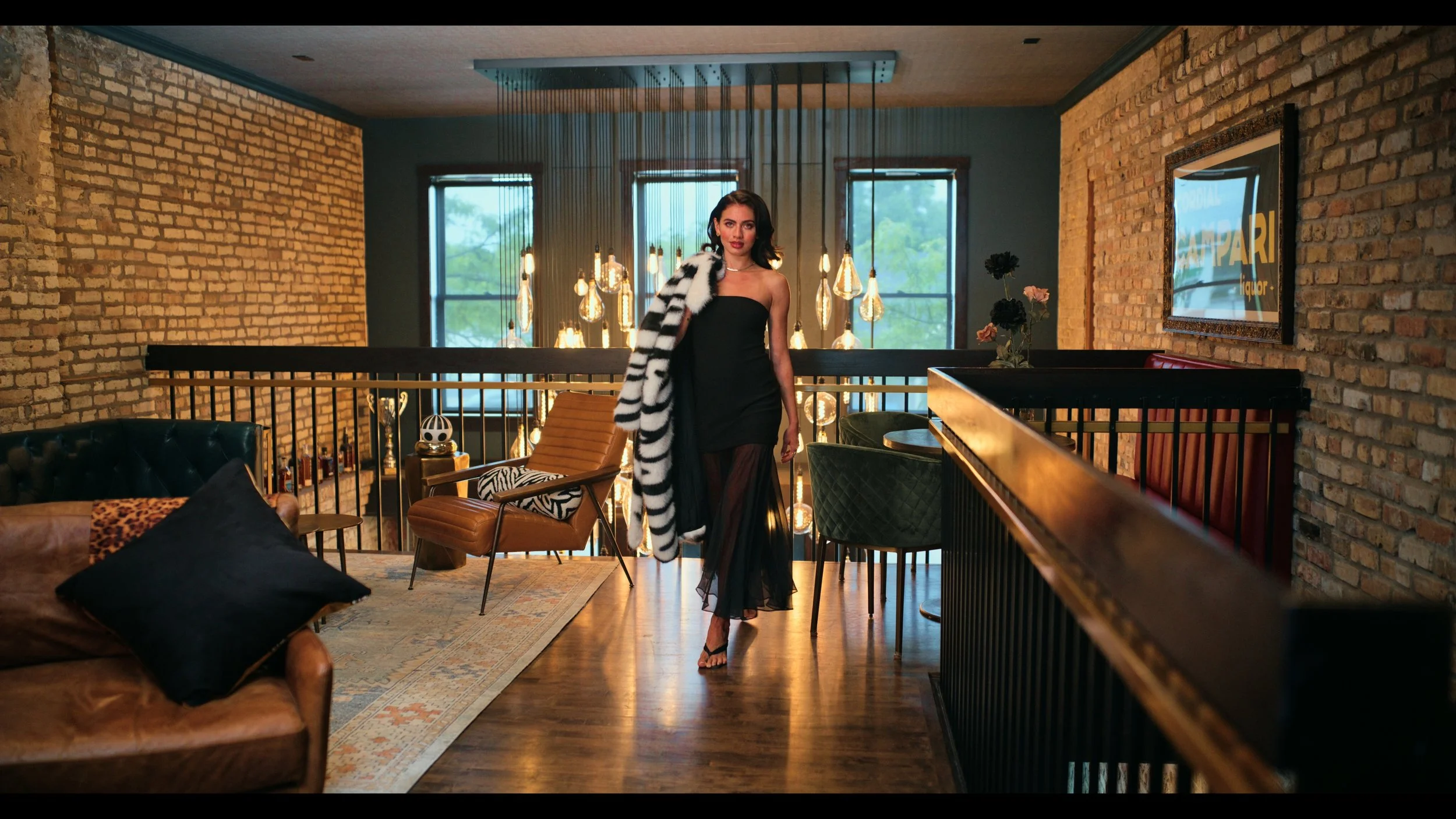 A woman in a black strapless dress with a striped coat draped over her shoulder, standing in a stylish, modern interior with exposed brick walls, large windows, and hanging pendant lights.