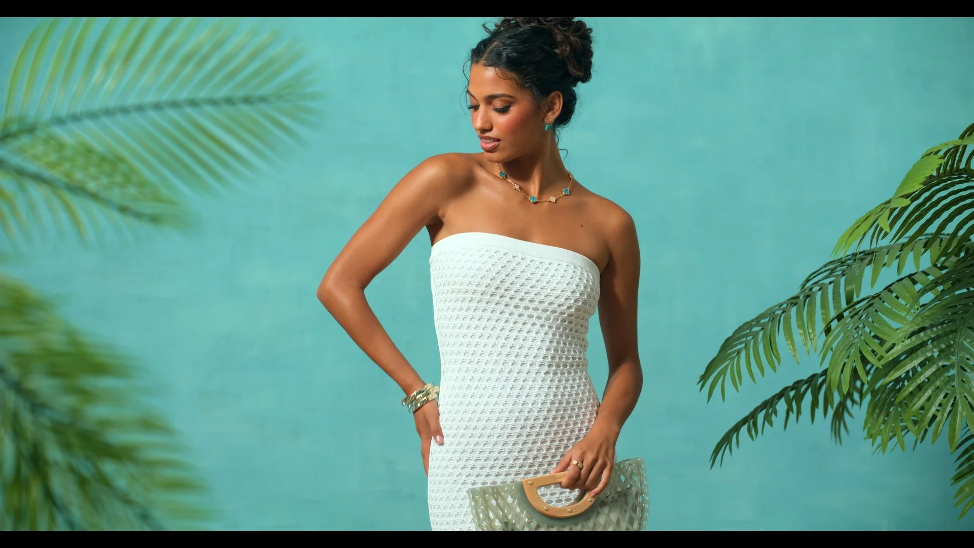 A woman in a white strapless textured dress standing against a turquoise background with green tropical foliage framing the sides. She is accessorized with jewelry and holding a stylish handbag.