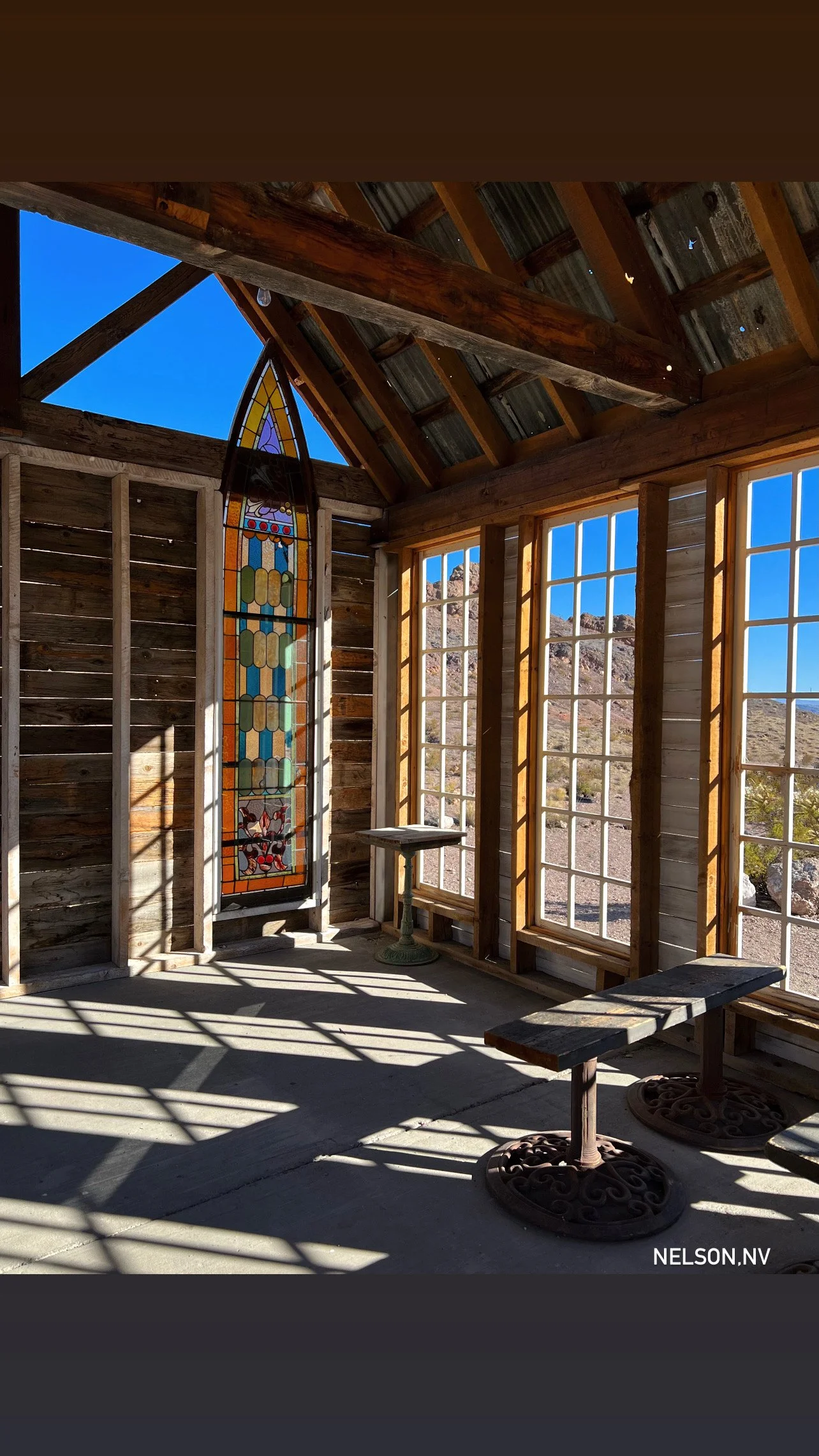 Interior of an unfinished wooden building with large windows, a stained glass window with a colorful design, and outdoor landscape visible through the windows and roof opening. Shadows cast by sunlight on the floor, wooden benches, and small table wi
