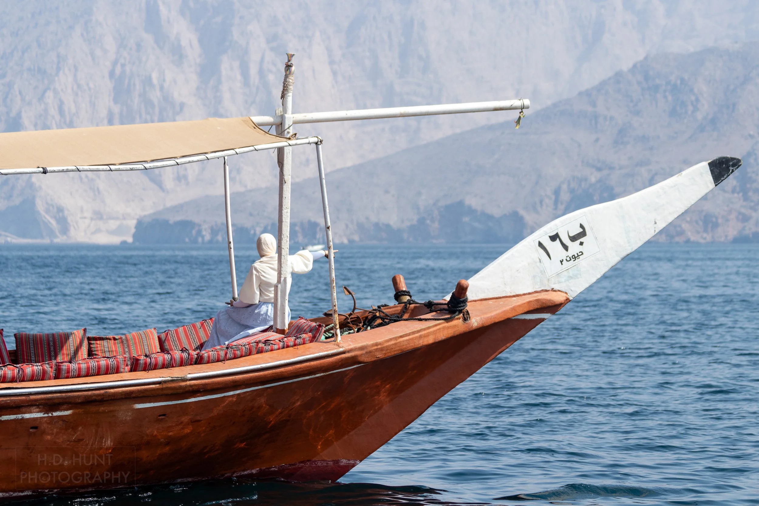 A woman points from the front of a dhow boat off into the distance, indicating a dolphin spotting off the Musandam Peninsula, Oman.