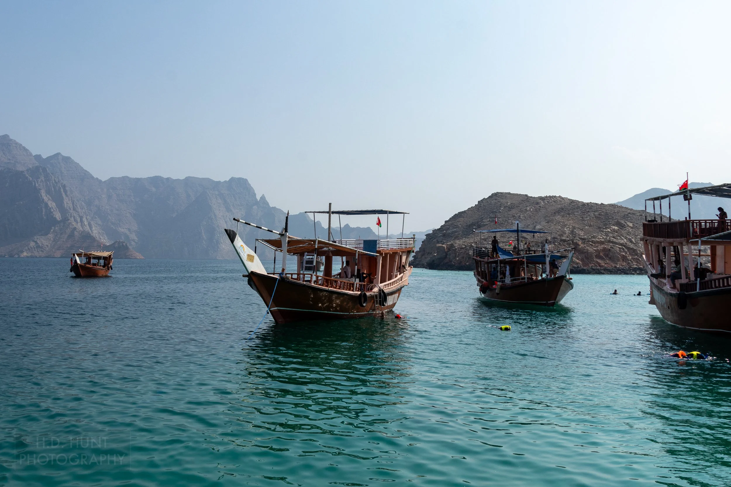 Four dhow boats are anchored in green-blue water while several visitors wearing brightly-colored lifejackets snorkel in the sea off the Musandam Peninsula, Oman.