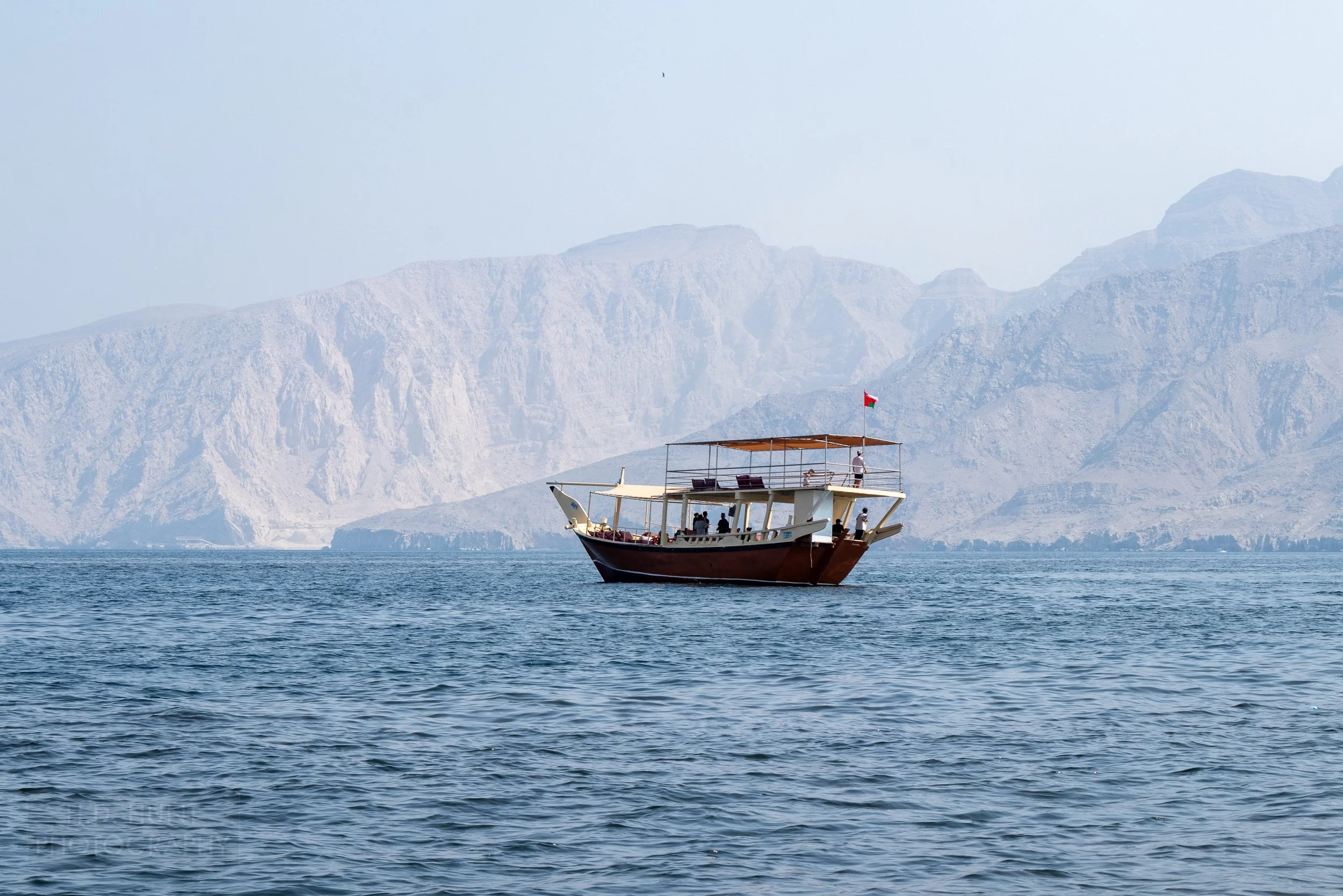 A dhow boat sits in the waters of the Musandam Peninsula, with prominent mountains in the background.