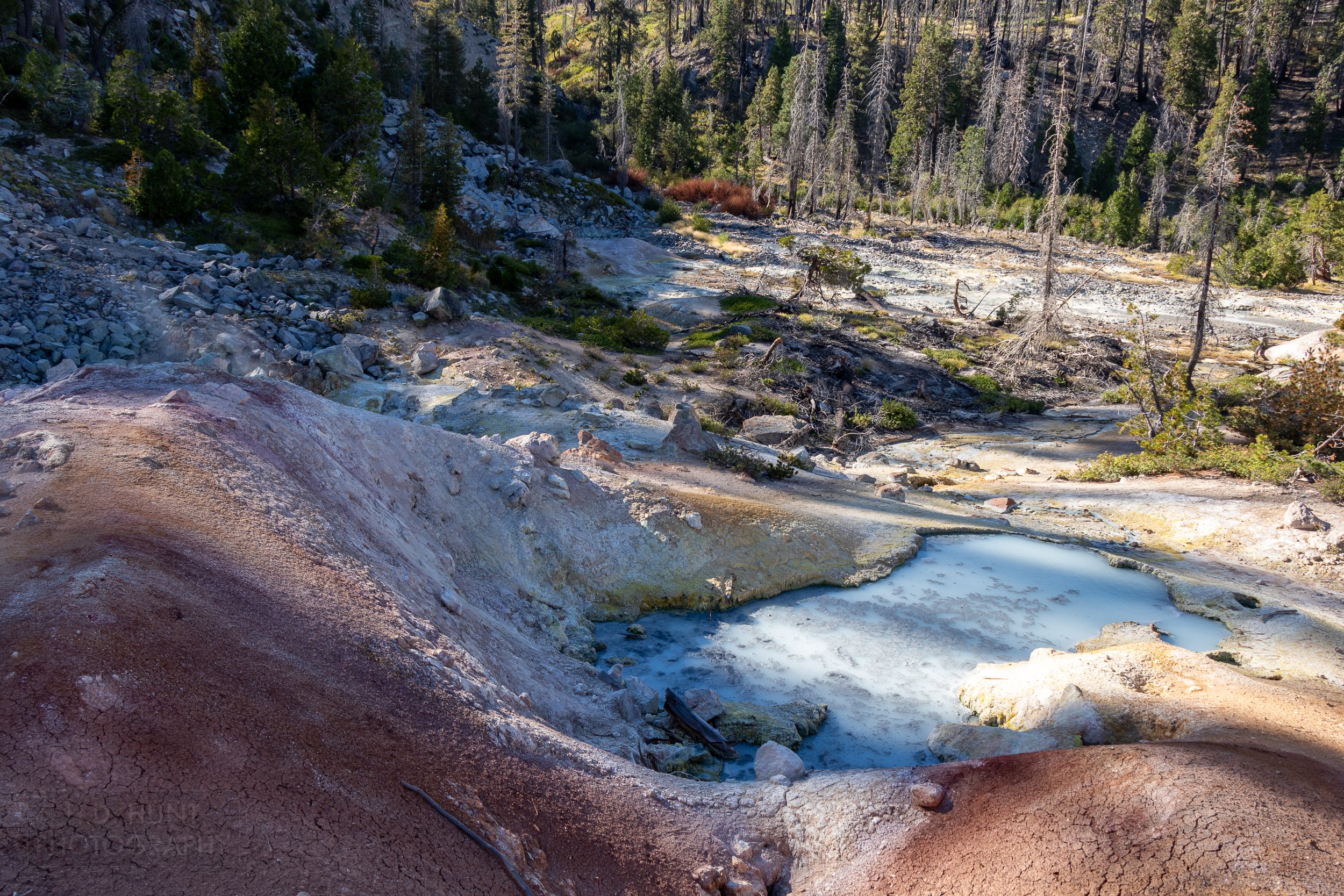 A pool of light blue and grey water sits on a barren landscape of red, orange, and white rocks, Devils Kitchen, Lassen Volcanic National Park, California, United States.