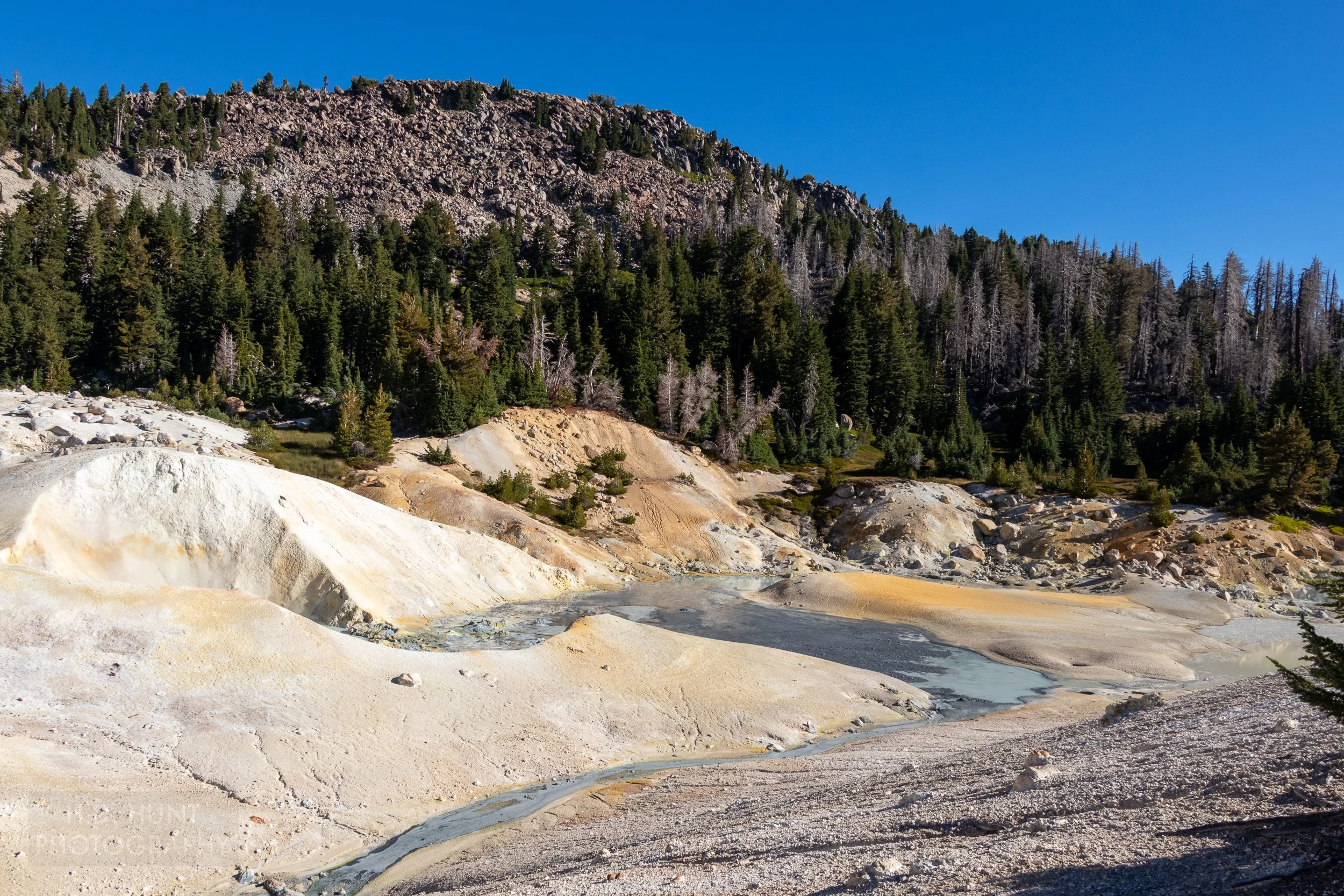 A stream of blue and grey water runs across white and tan rock at Bumpass Hell, Lassen Volcanic National Park, California, United States.