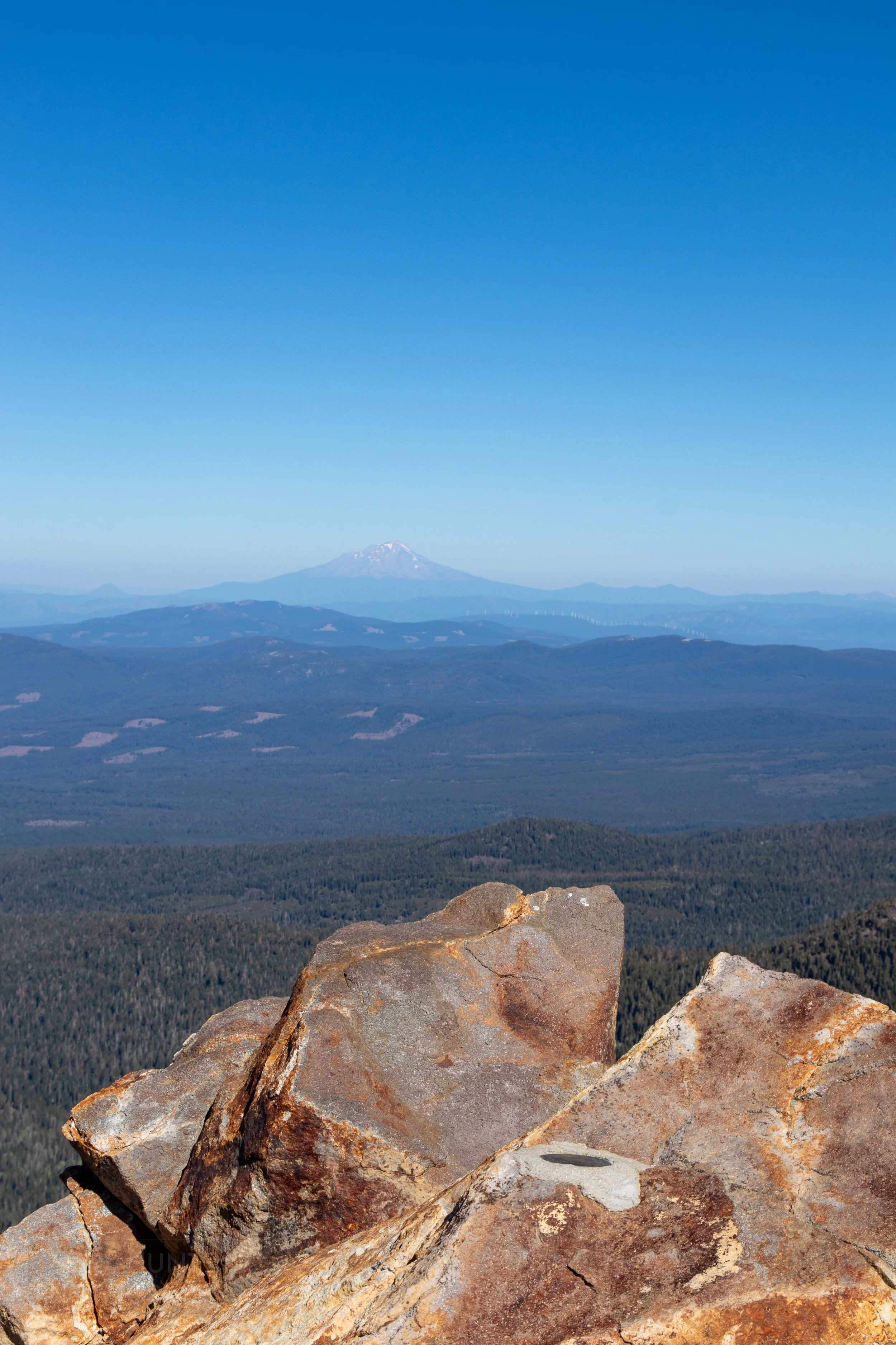 Mount Shasta is seen far in the distance from the top of Brokeoff Mountain, Lassen Volcanic National Park, California, United States; a round metal surveying marker is seen in the foreground atop a brown and red rocky outcropping.
