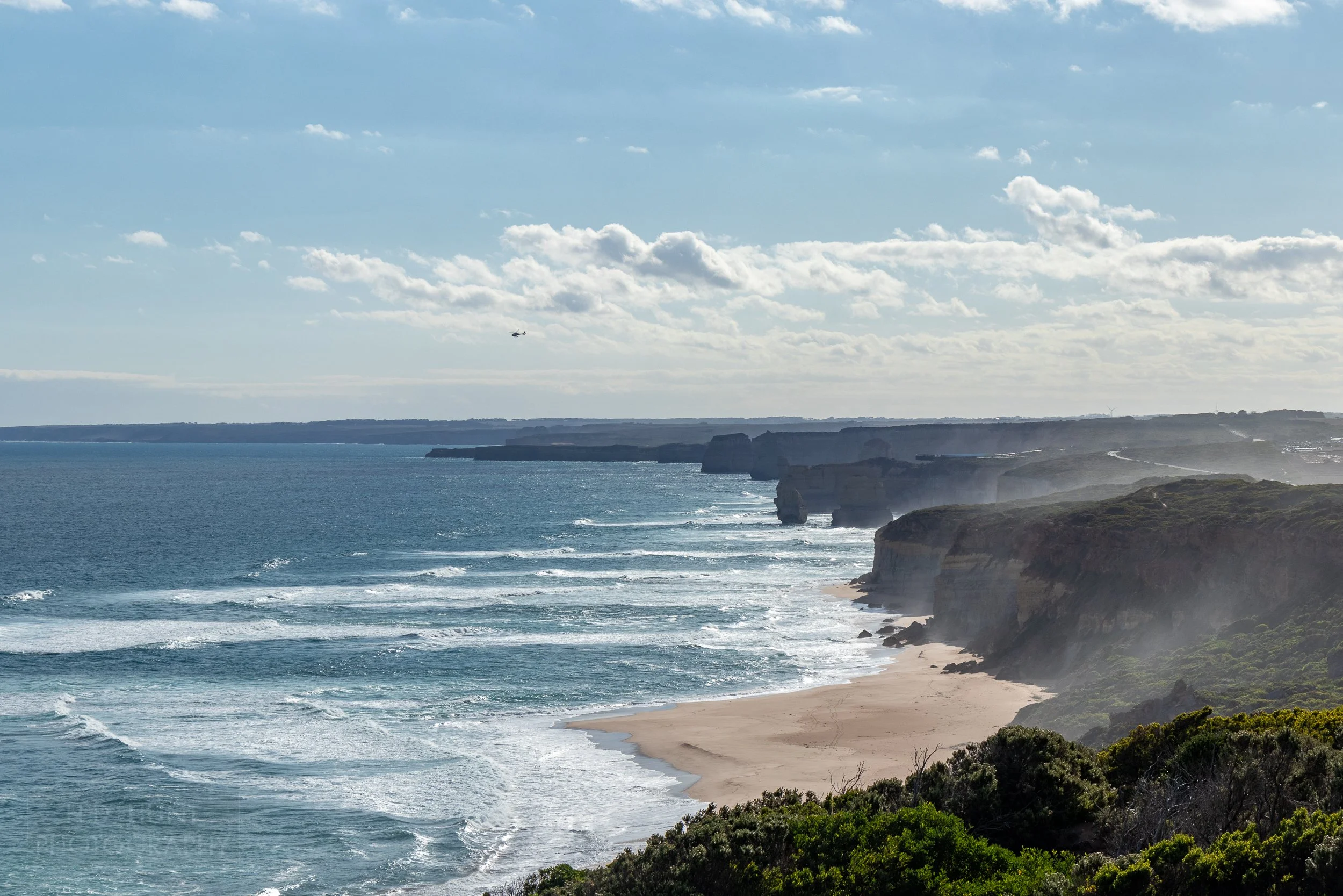 The towers of The Twelve Apostles rock formation can be seen along the coast while a helicopter flies overhead in the distance, The Great Ocean Walk, Victoria, Australia.
