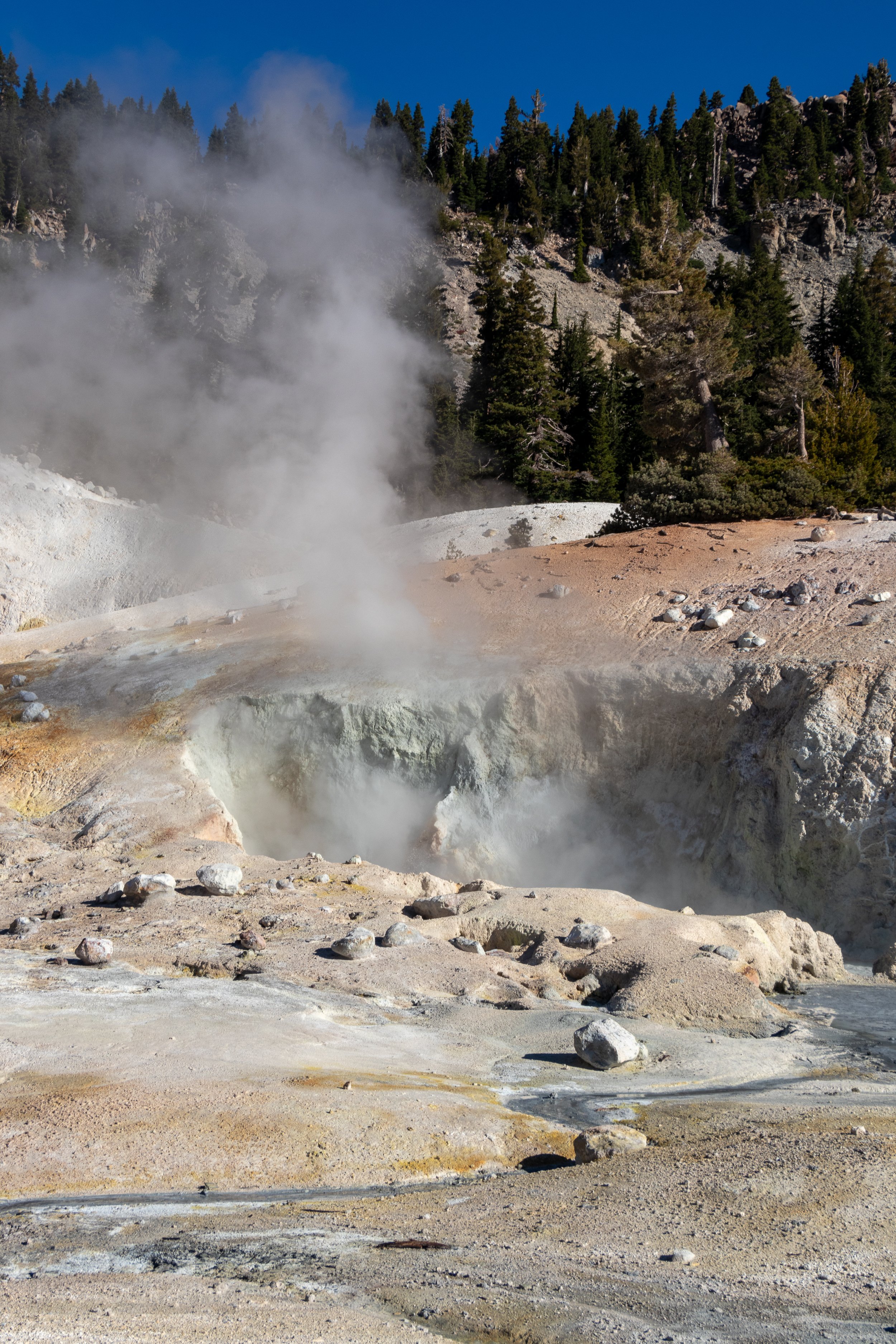 Steam rises from a white, grey, and tan hole of earth at Bumpass Hell, Lassen Volcanic National Park, California, United States.