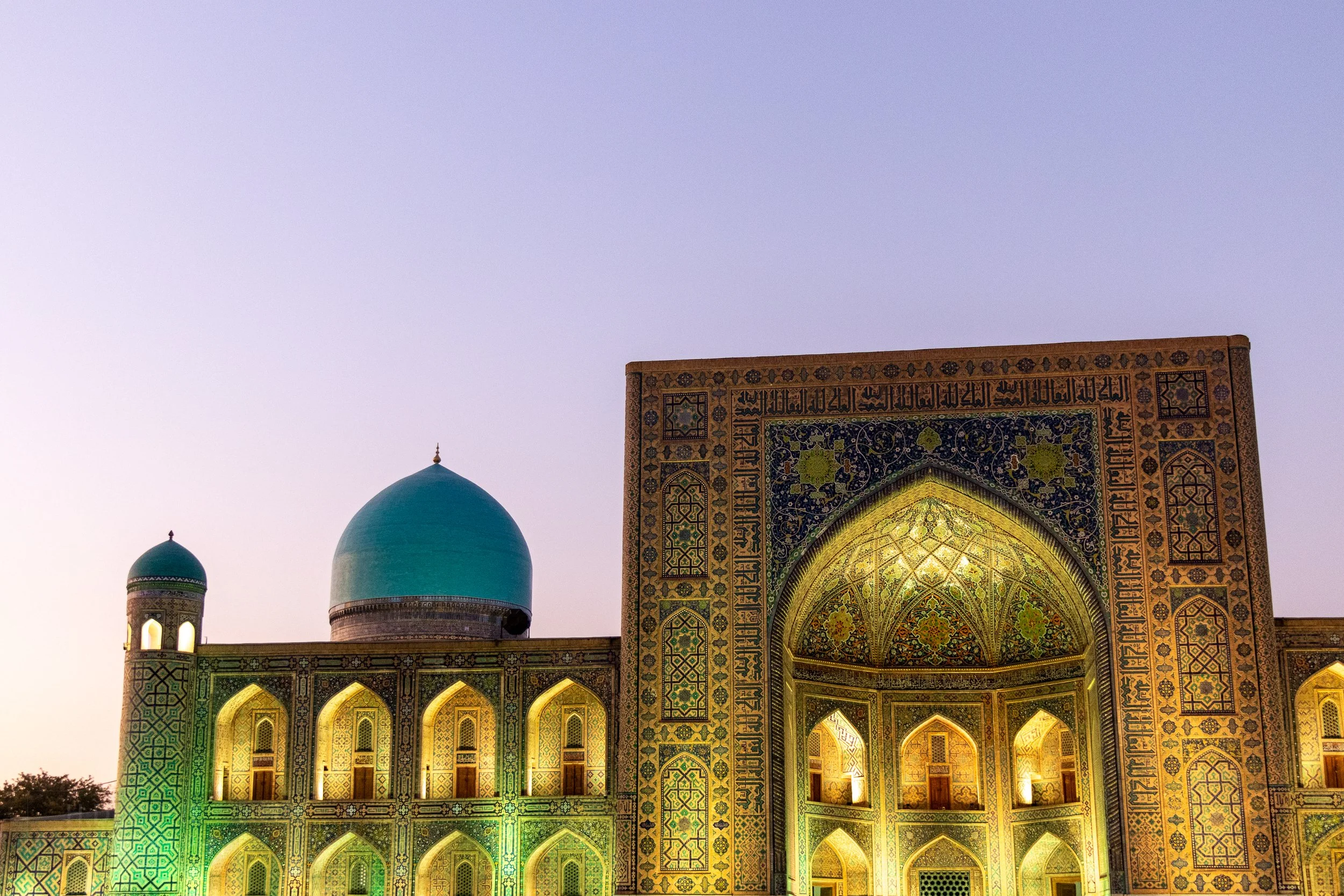 Exterior of the Tilya Kori Madrasa at sunset, featuring a large blue dome and an ornately-tiled archway, Registan, Samarkand, Uzbekistan.
