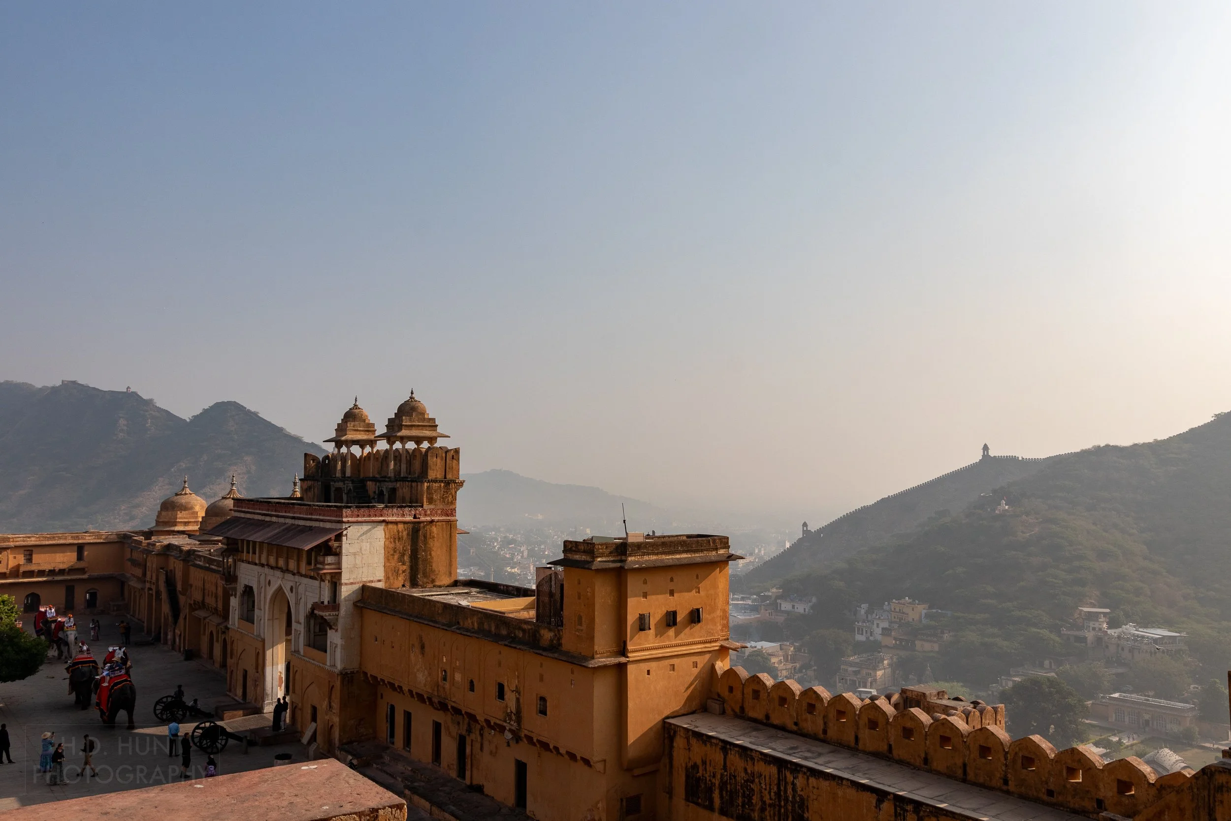 A large stone gate stands above a broad valley and beside a courtyard featuring elephants with visitors on their backs, Amber Fort, Amer, India.