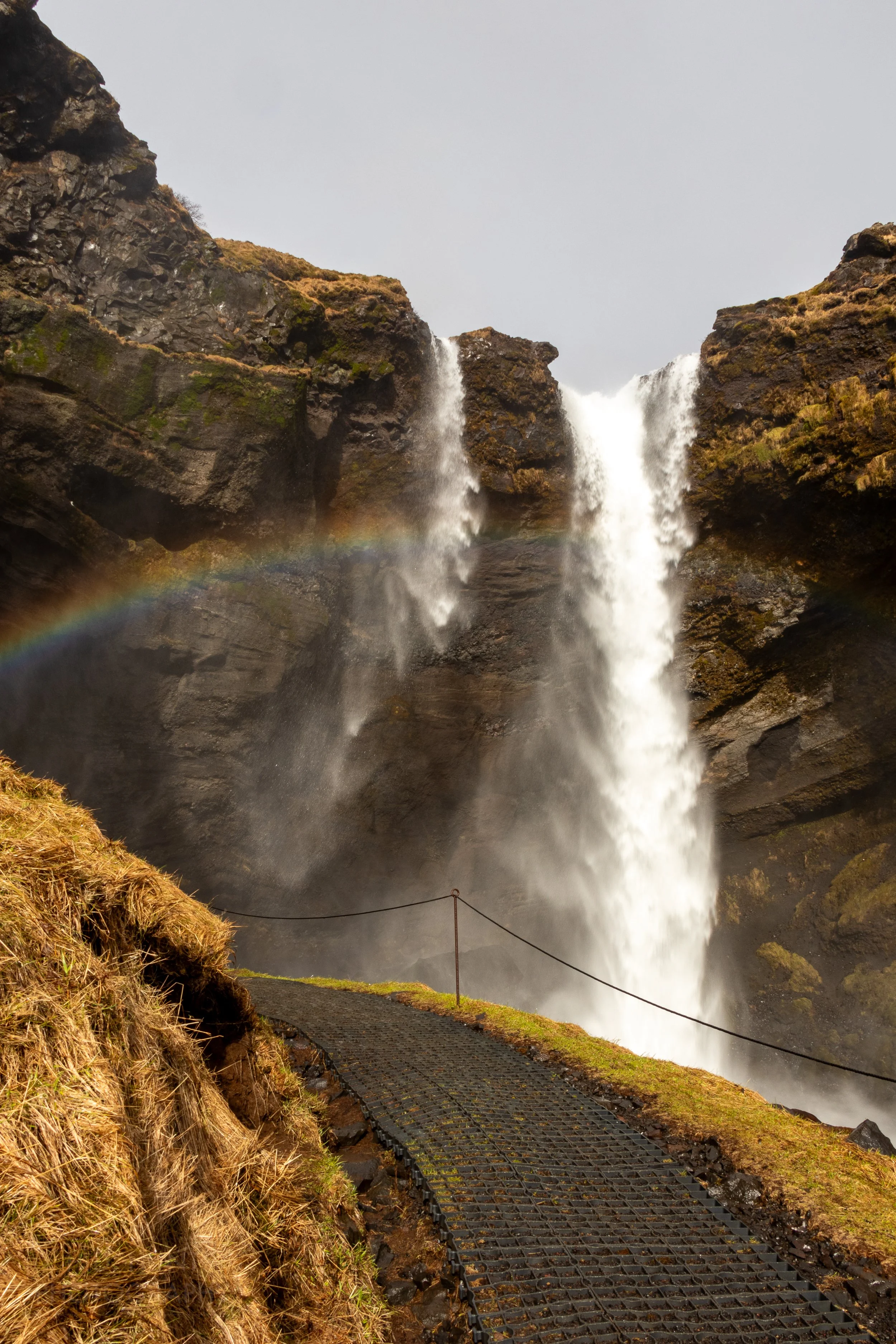 A walking path leads to a waterfall with two drops over a dark rock cliff with a rainbow in its foreground, Kvernufoss, Iceland.