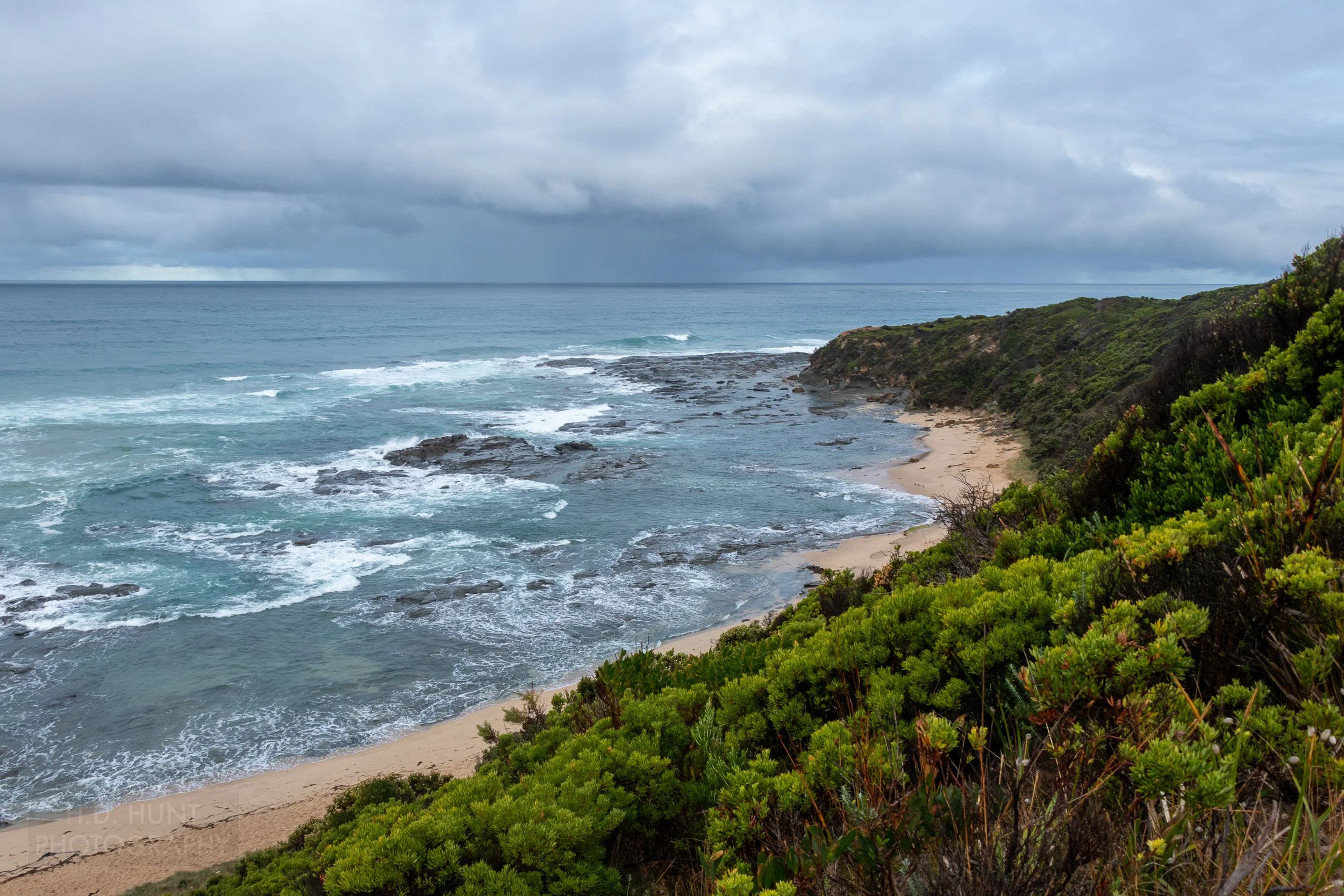 Waves crash along rocks beneath small bush-covered hills along The Great Ocean Walk, Victoria, Australia.