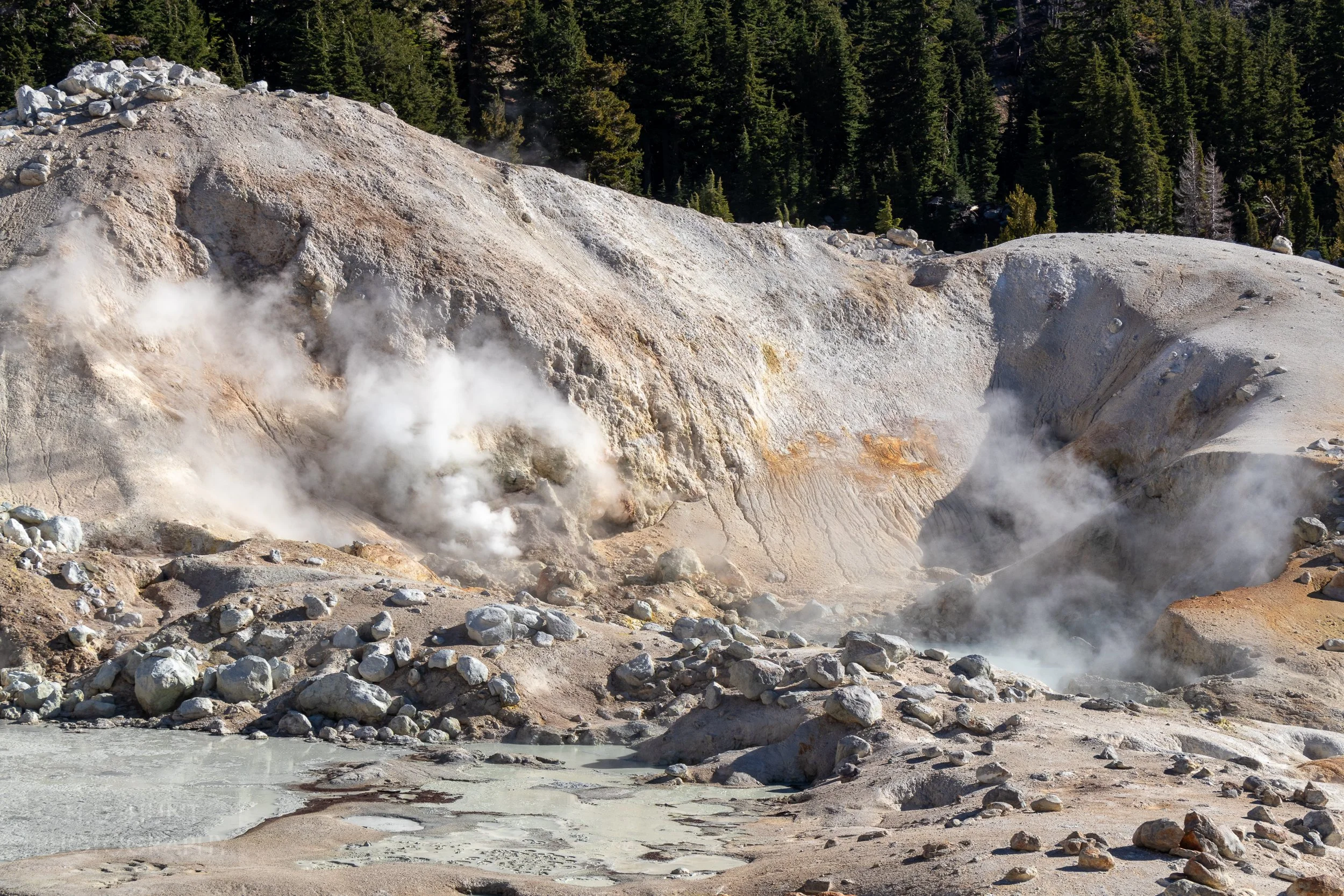 Steam rises from multiple earth holes in front of a hill of white and tan rock, Bumpass Hell, Lassen Volcanic National Park, California, United States.