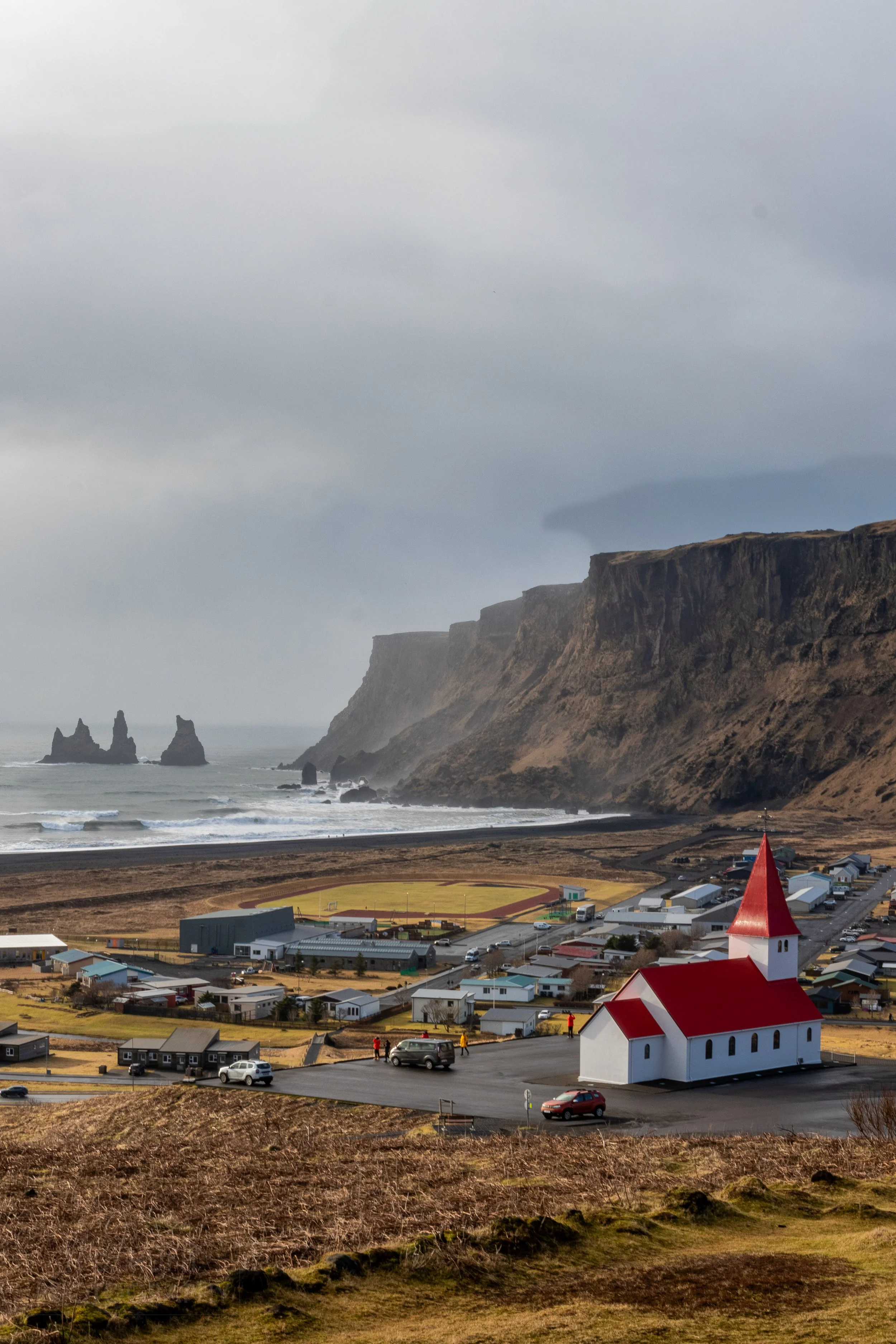 The Vík í Mýrdal Church is seen, with the village of Vík í Mýrdal in the mid-ground, and a black sand beach beneath tall cliffs in the background, Vík í Mýrdal, Iceland.