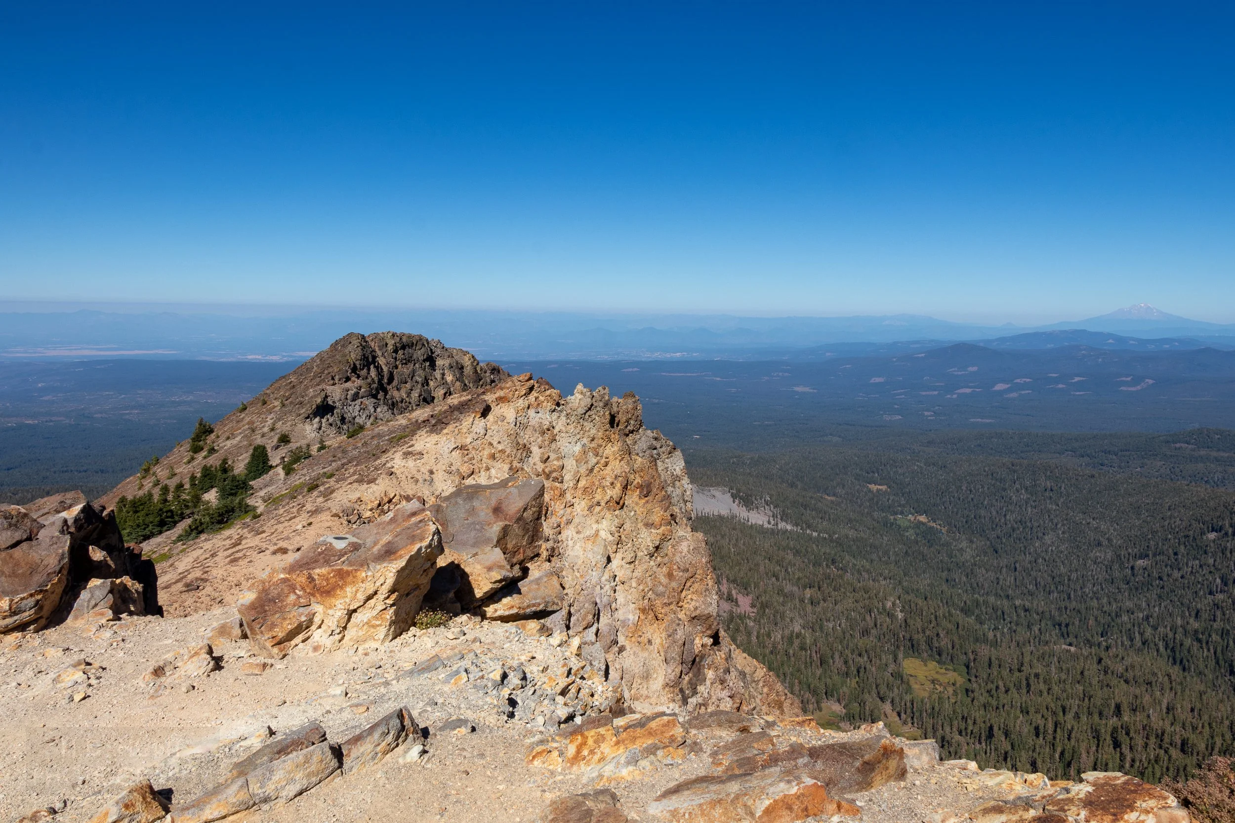 A rocky outcropping at the peak of Brokeoff Mountain is seen, with trees stretching far into the distance in the background, Lassen Volcanic National Park, California, United States.