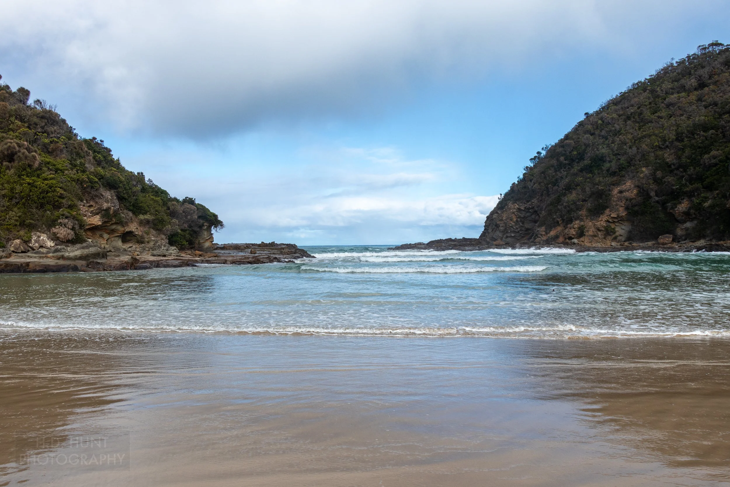 Waves splash into a small cove between two green plant-covered hills at Parker Inlet along The Great Ocean Walk, Victoria, Australia.