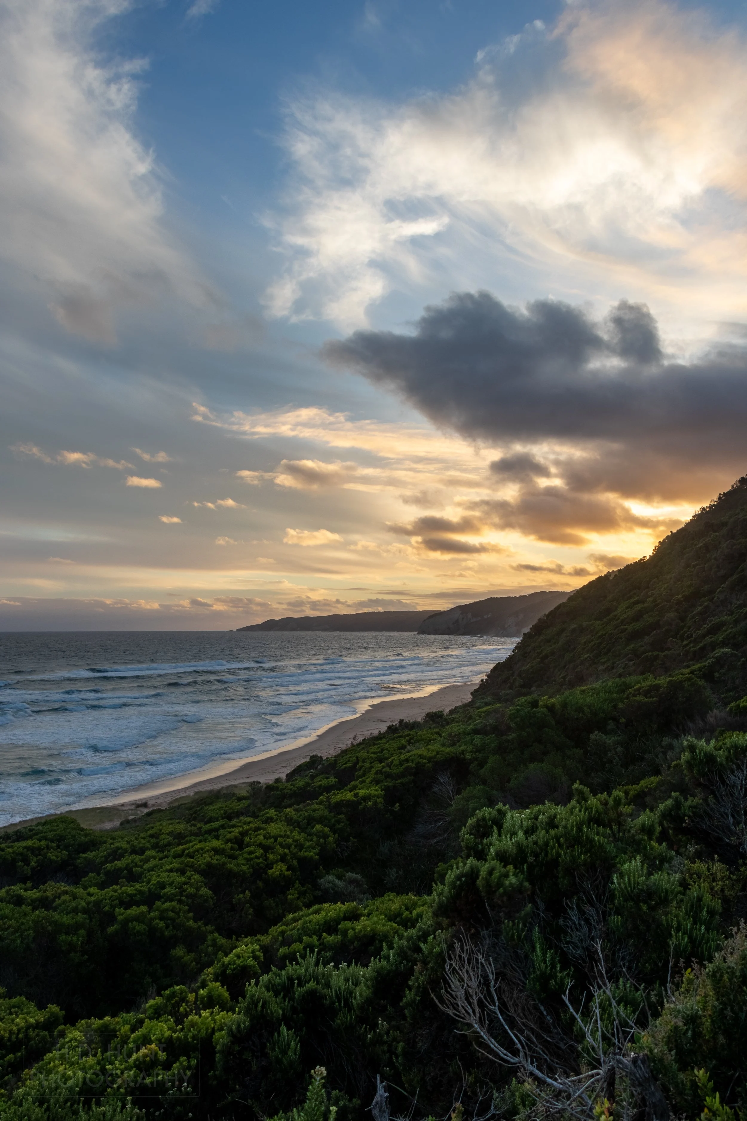 Sunset punches a hole through clouds above green shrub-covered cliffs near Johanna, Victoria, Australia.