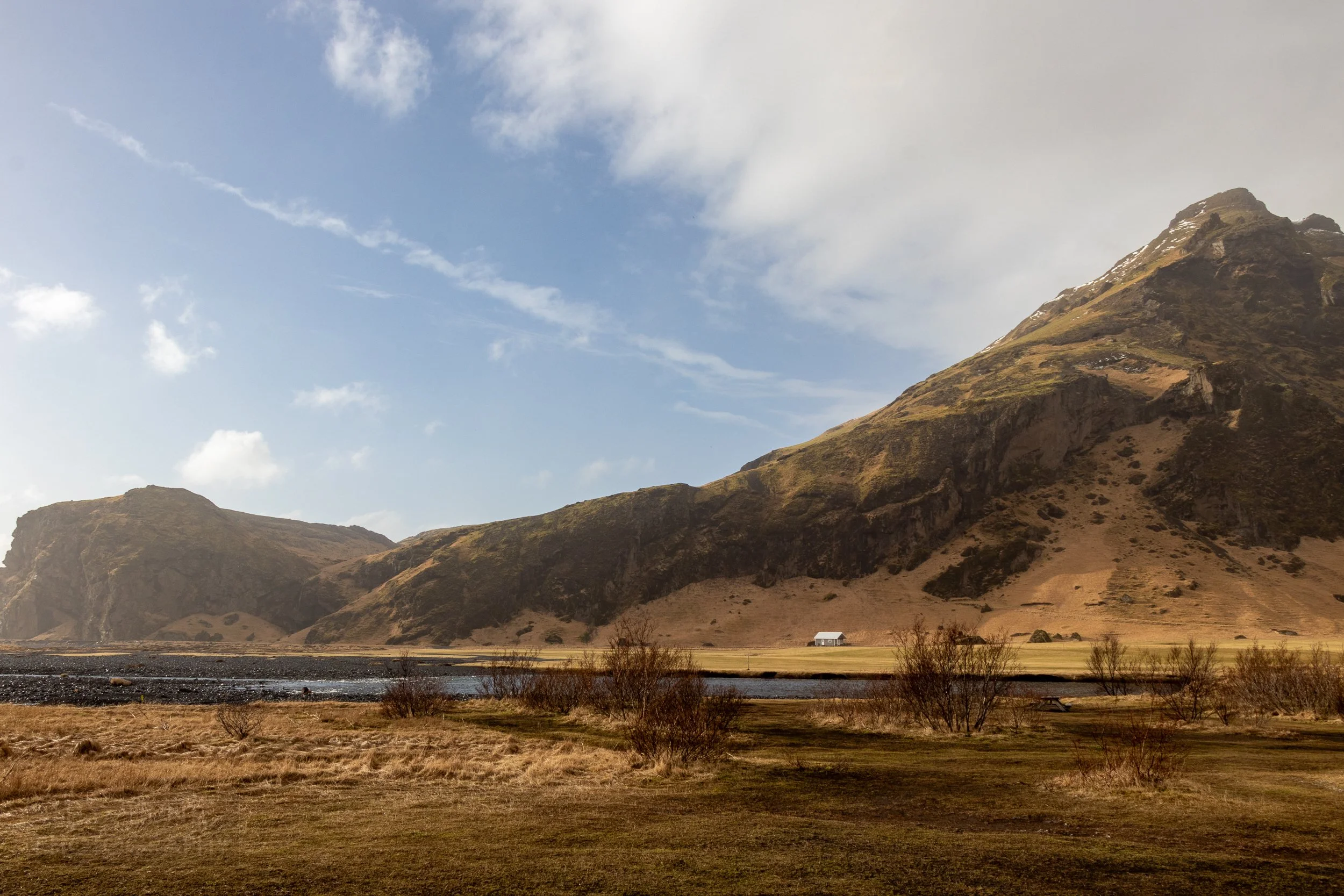 A mountain rises behind a river and a grass field near Skógafoss, Iceland.