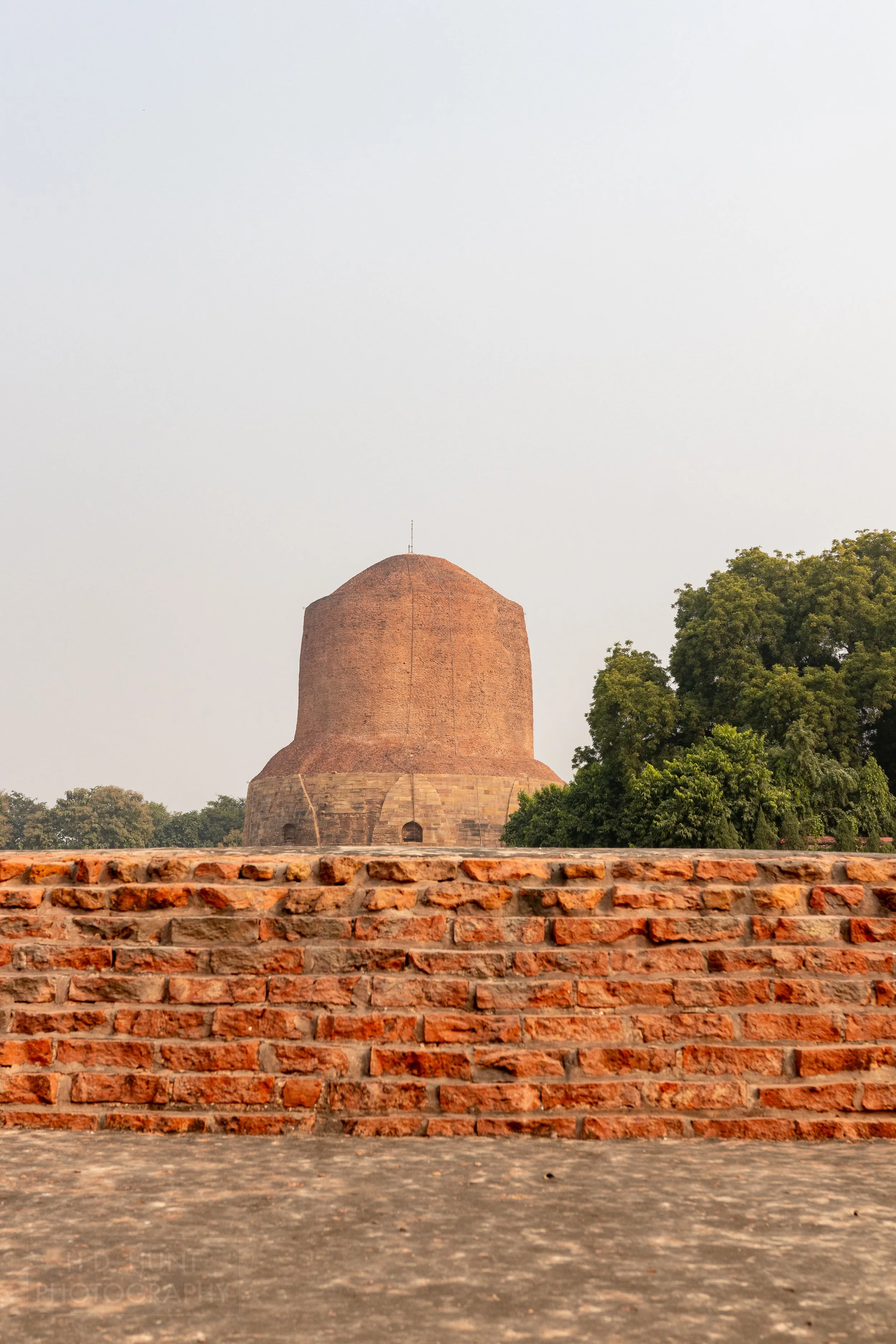 A large stone brick tower rises above a small brick wall, Sarnath, India.