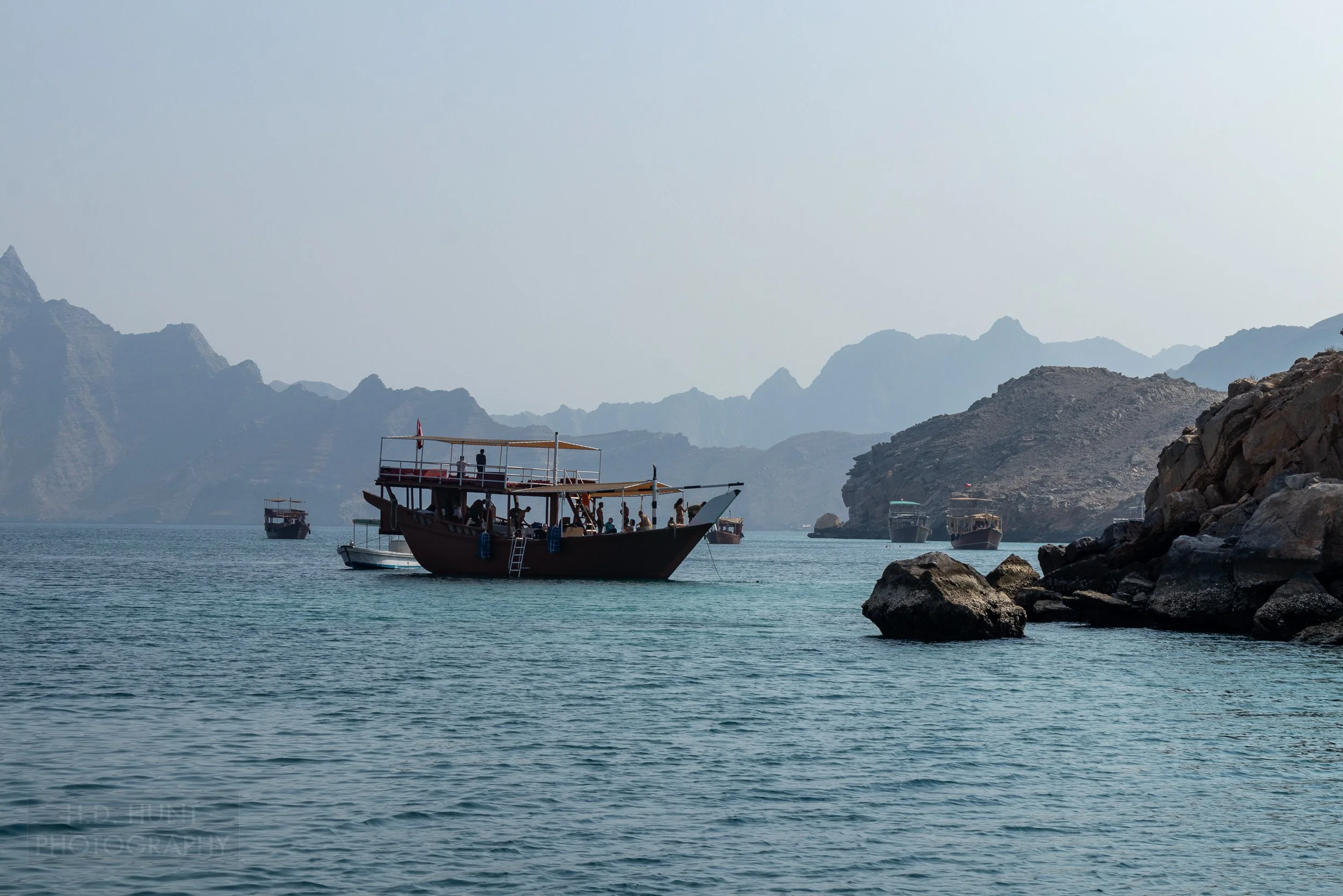 Several boats are anchored off a rocky outcrop and the mountains of the Musandam Peninsula, Oman, rise in the background.
