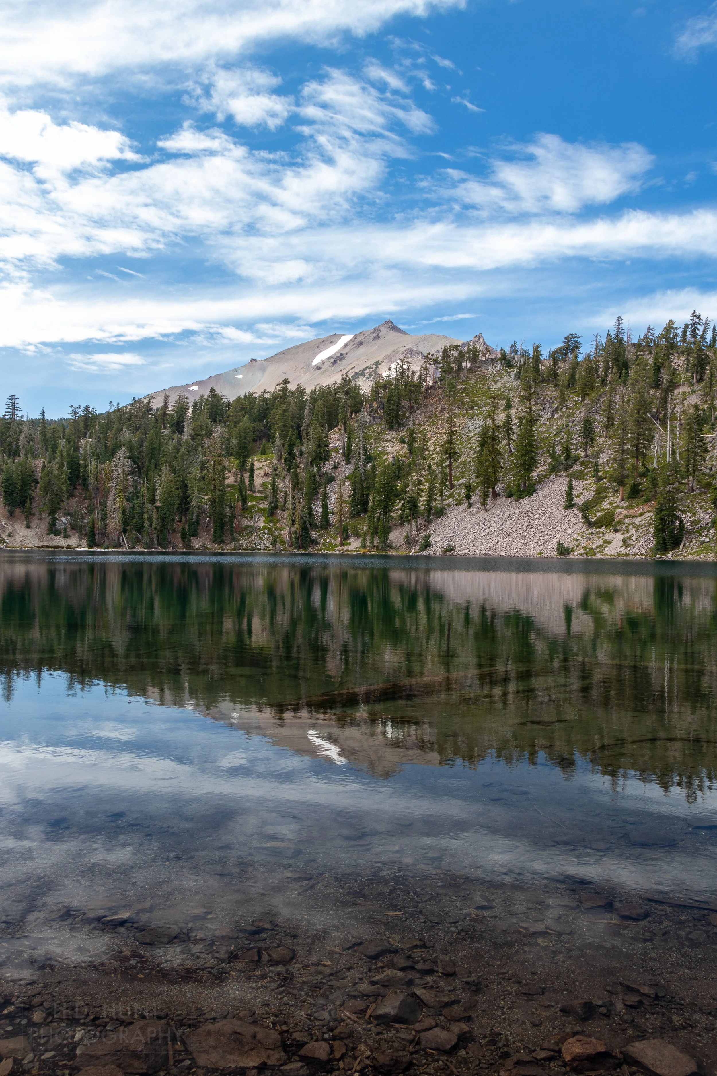 A tall mountain - Lassen Peak - is seen behind a clear blue lake, Lassen Volcanic National Park, California, United States.