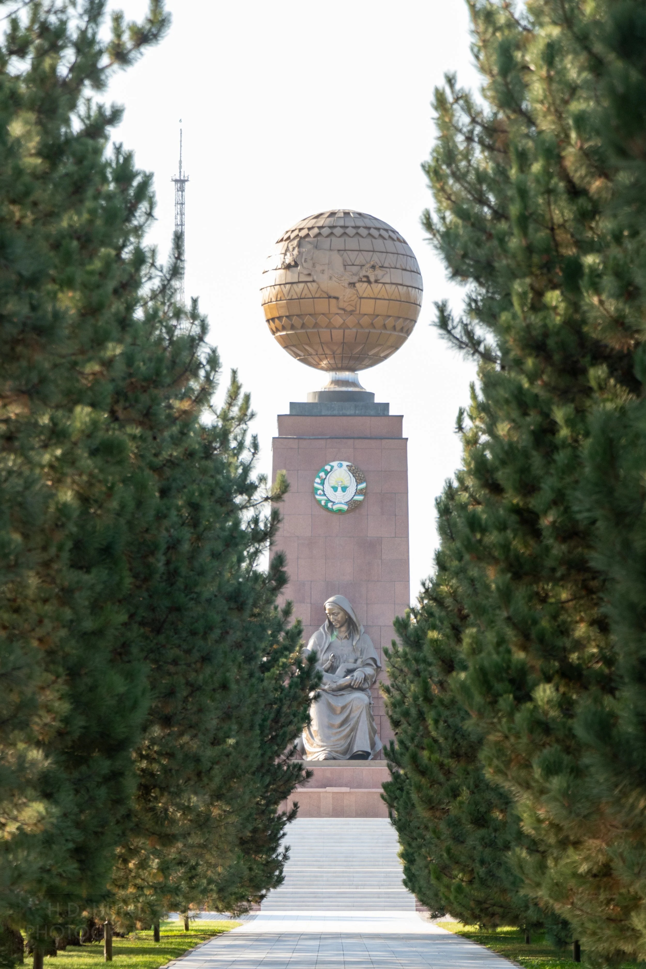 A red stone column with a copper-colored globe featuring Uzbekistan rises above a metal statue of a woman holding a baby in Tashkent, Uzbekistan.