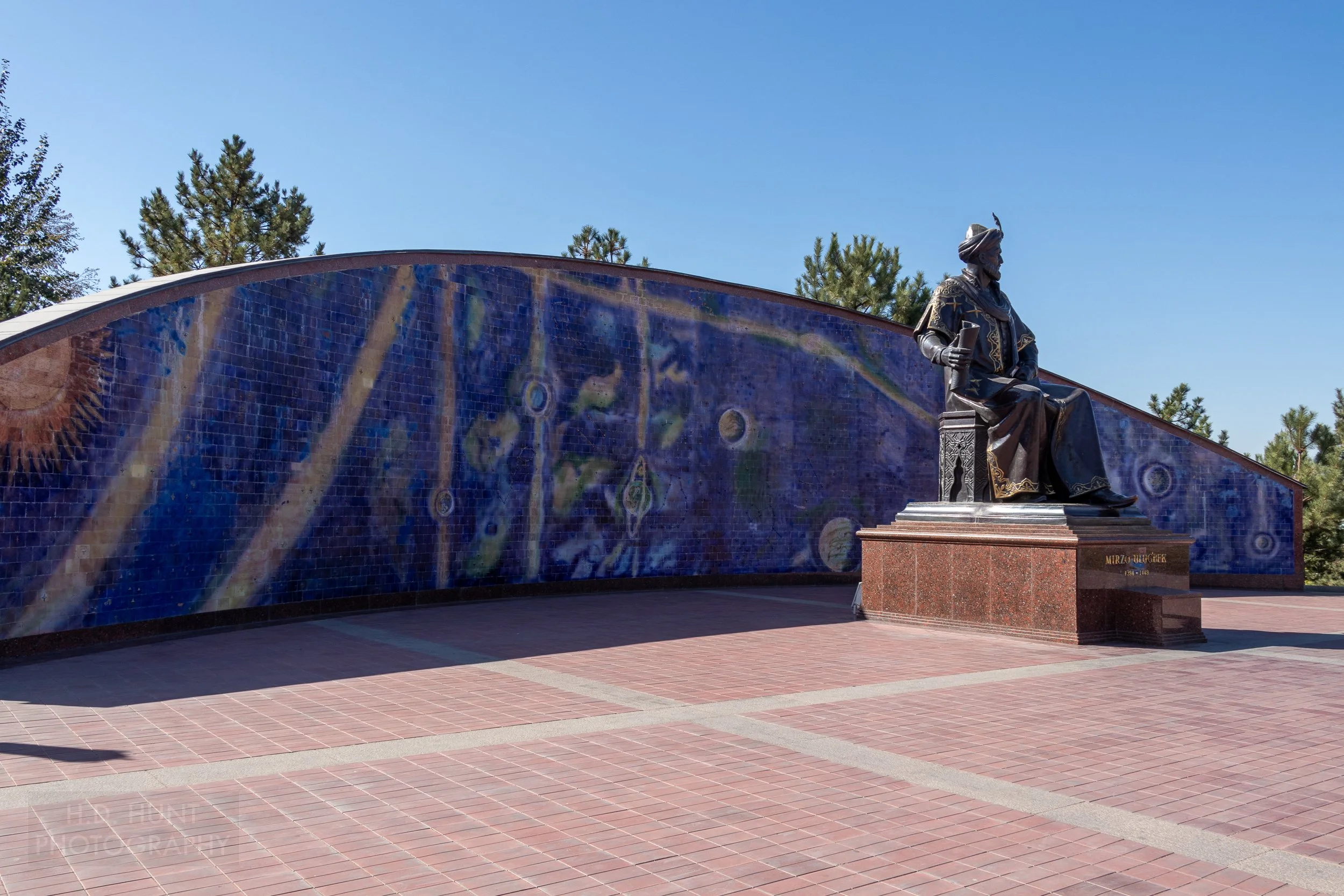 A large statue of a seated Ulugh Beg sits in a courtyard, behind which is a large stone wall featuring predominantly blue paint depicting the solar system, Samarkand, Uzbekistan.
