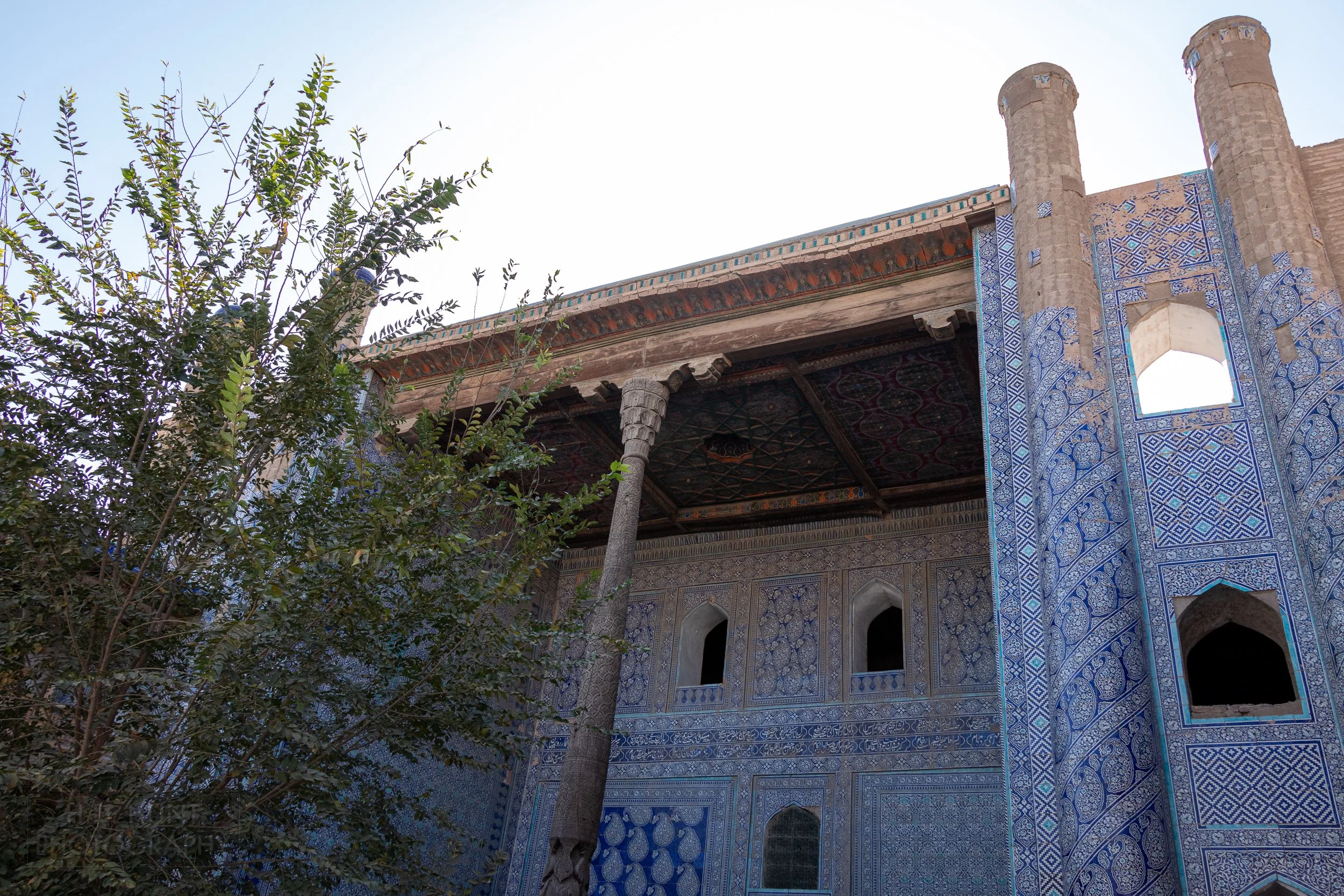 A wooden pillar holds up a portico adorned with intricate blue and white tile work at the Toshhovli Palace, Khiva, Uzbekistan.