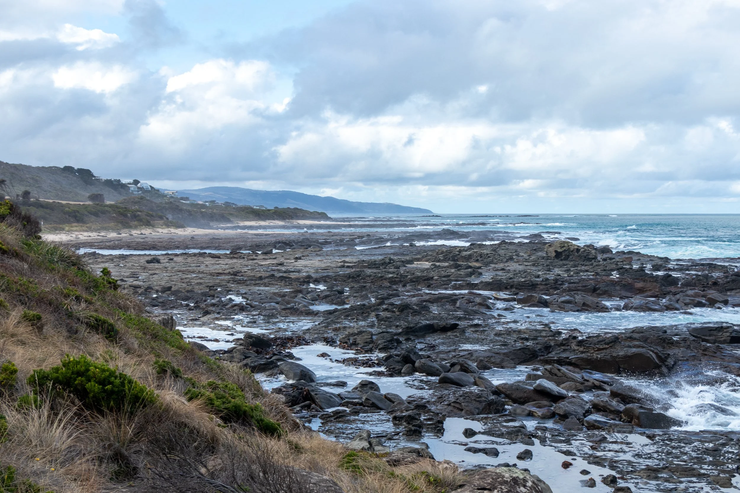 Waves crash into rocks along the Bass Strait beneath a green grass-covered cliff along The Great Ocean Walk, Victoria, Australia.