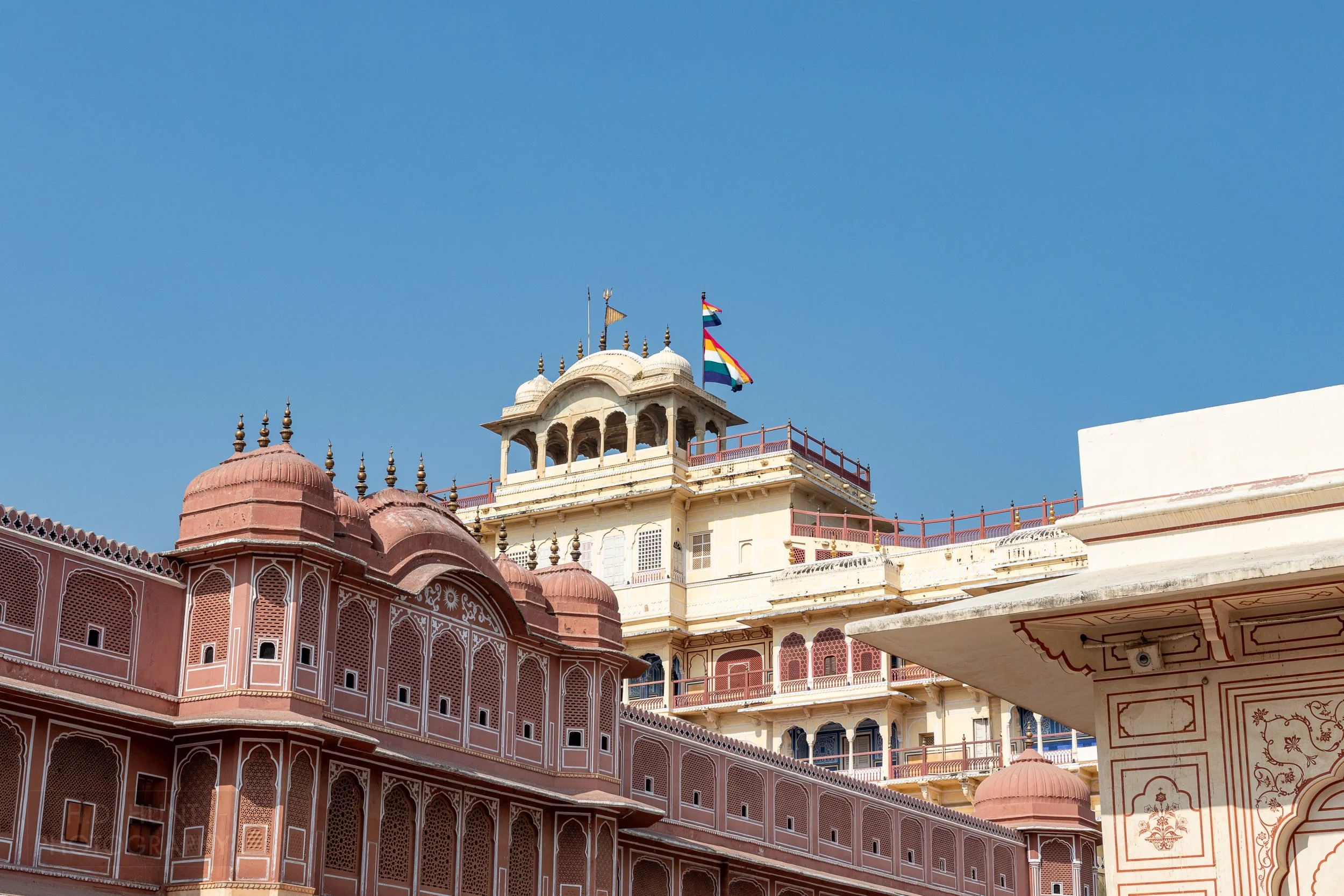 A large white building with multi-colored flags sits above a lower pink colored stone building, City Palace, Jaipur, India.