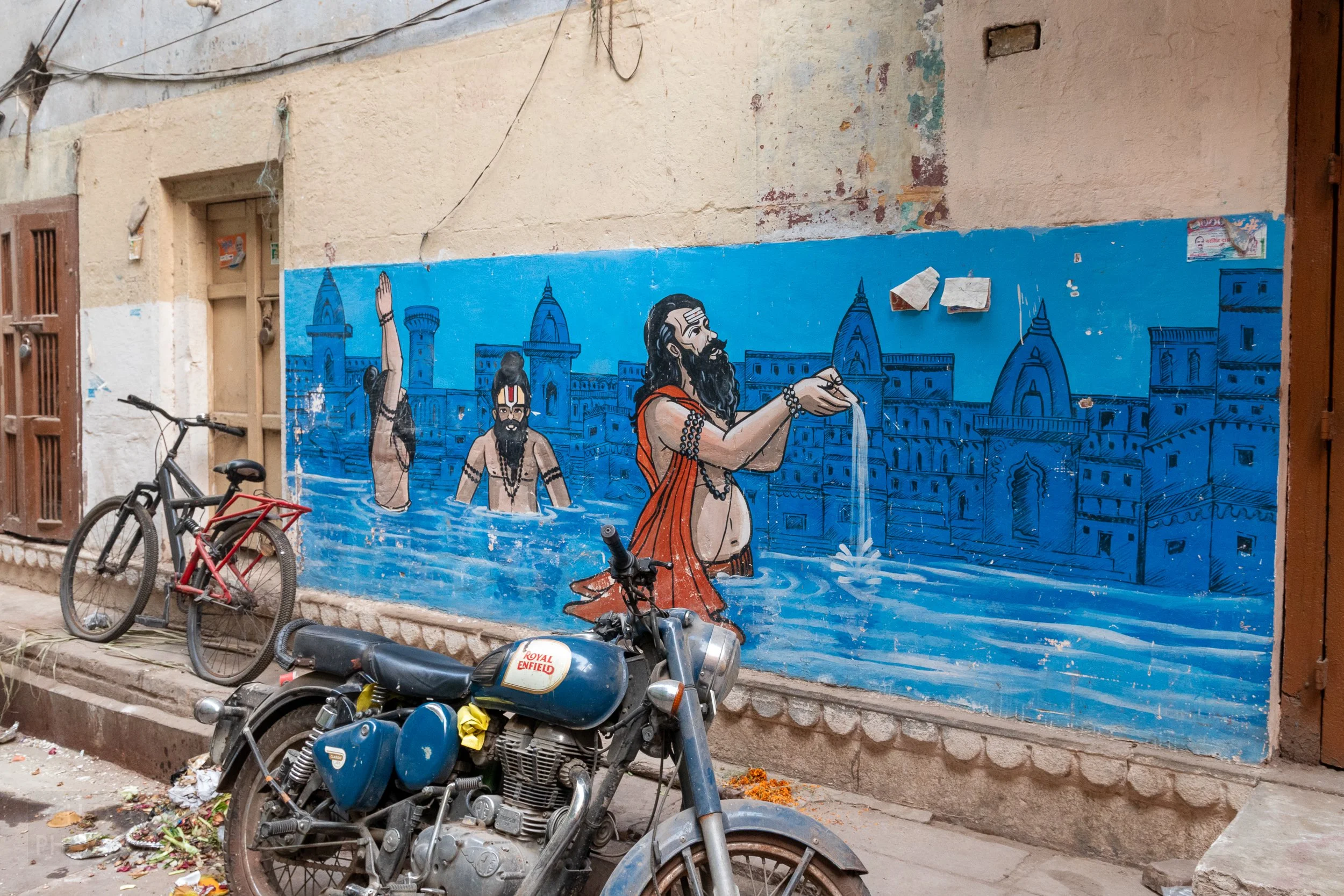 A painted blue mural depicting a man wearing an orange wrap standing in water appears in front of a motorcycle in an alley in Varanasi, India.