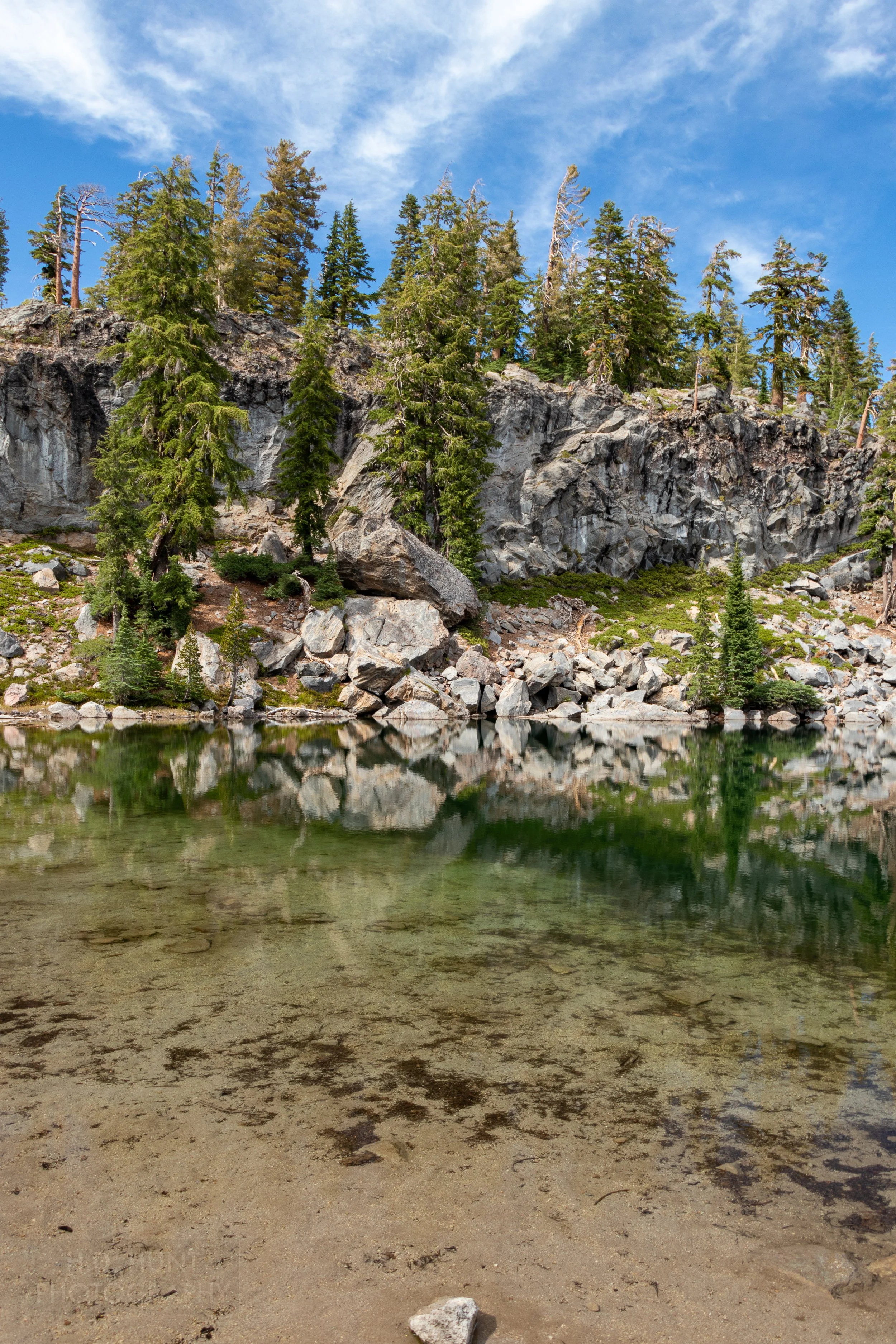 A green lake reflects a nearby rocky cliff and trees, Lassen Volcanic National Park, California, United States.