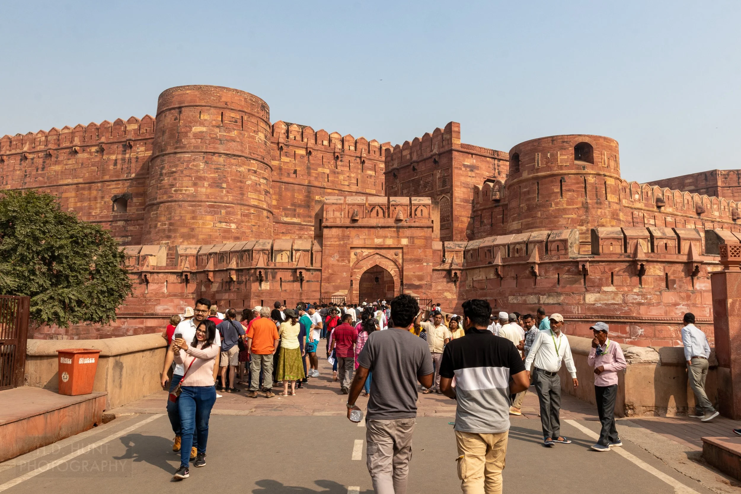 The large exterior fortress walls of Agra Fort are seen behind dozens of visitors who stand on a stone bridge, Agra, India.