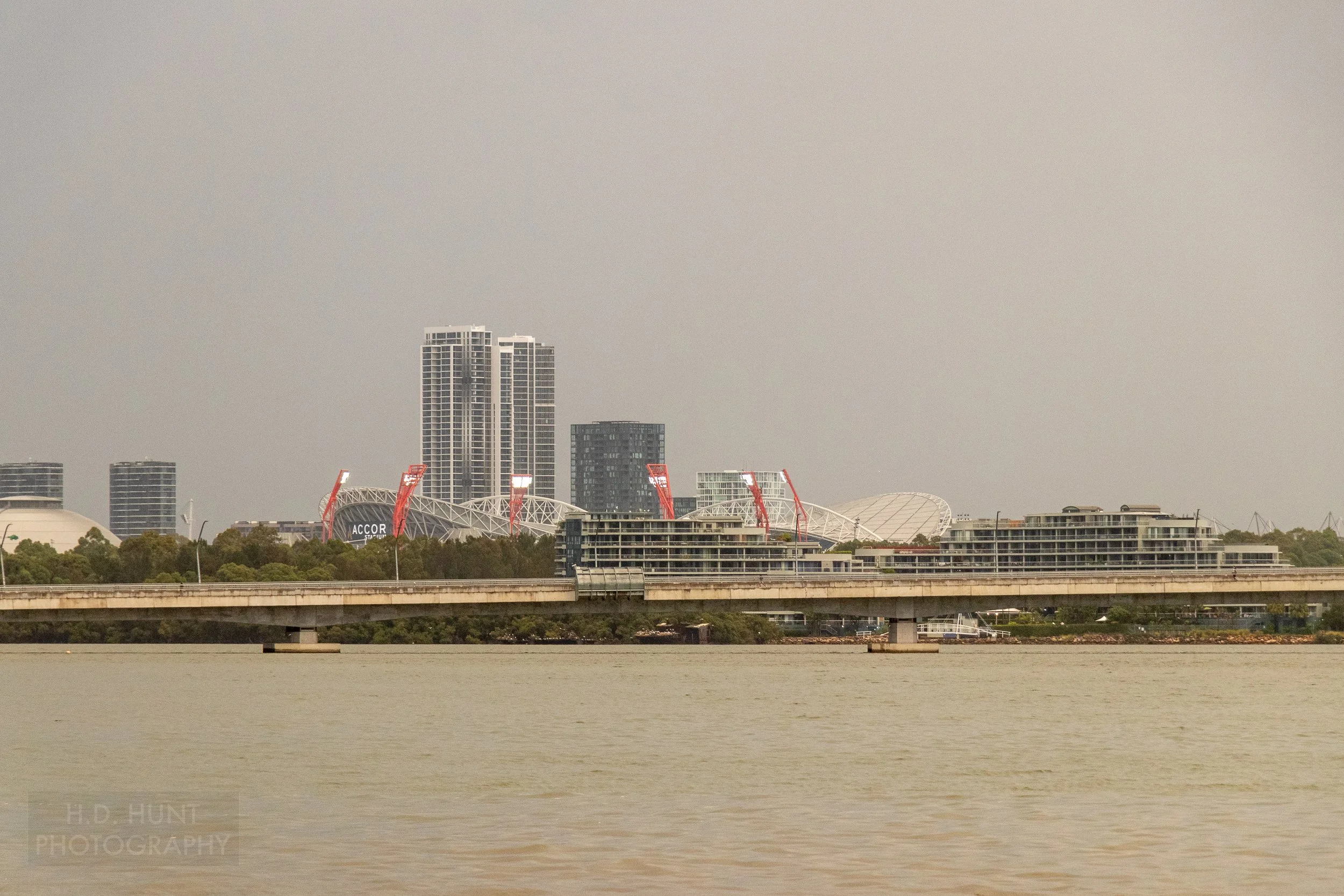 A large stadium and skyscrapers are seen on an overcast day from the Parramatta River, Australia.