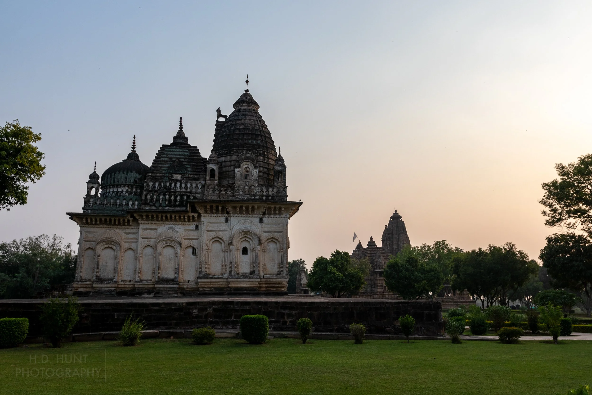 The Pratapeshwar Temple stands in the foreground as the sun sets behind the Khajuraho Group of Monuments, India.