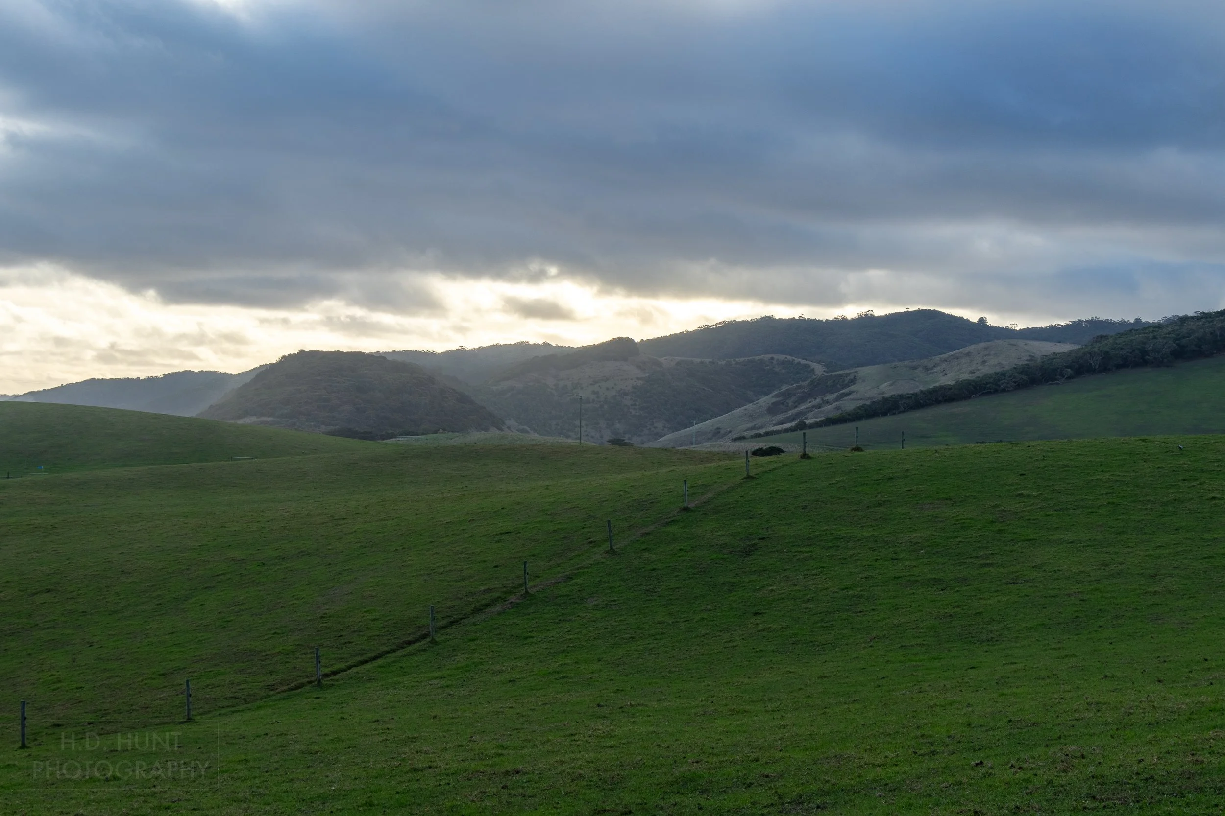 The sun peeks from behind thick, dark clouds above thick green grass in Johanna, Victoria, Australia.