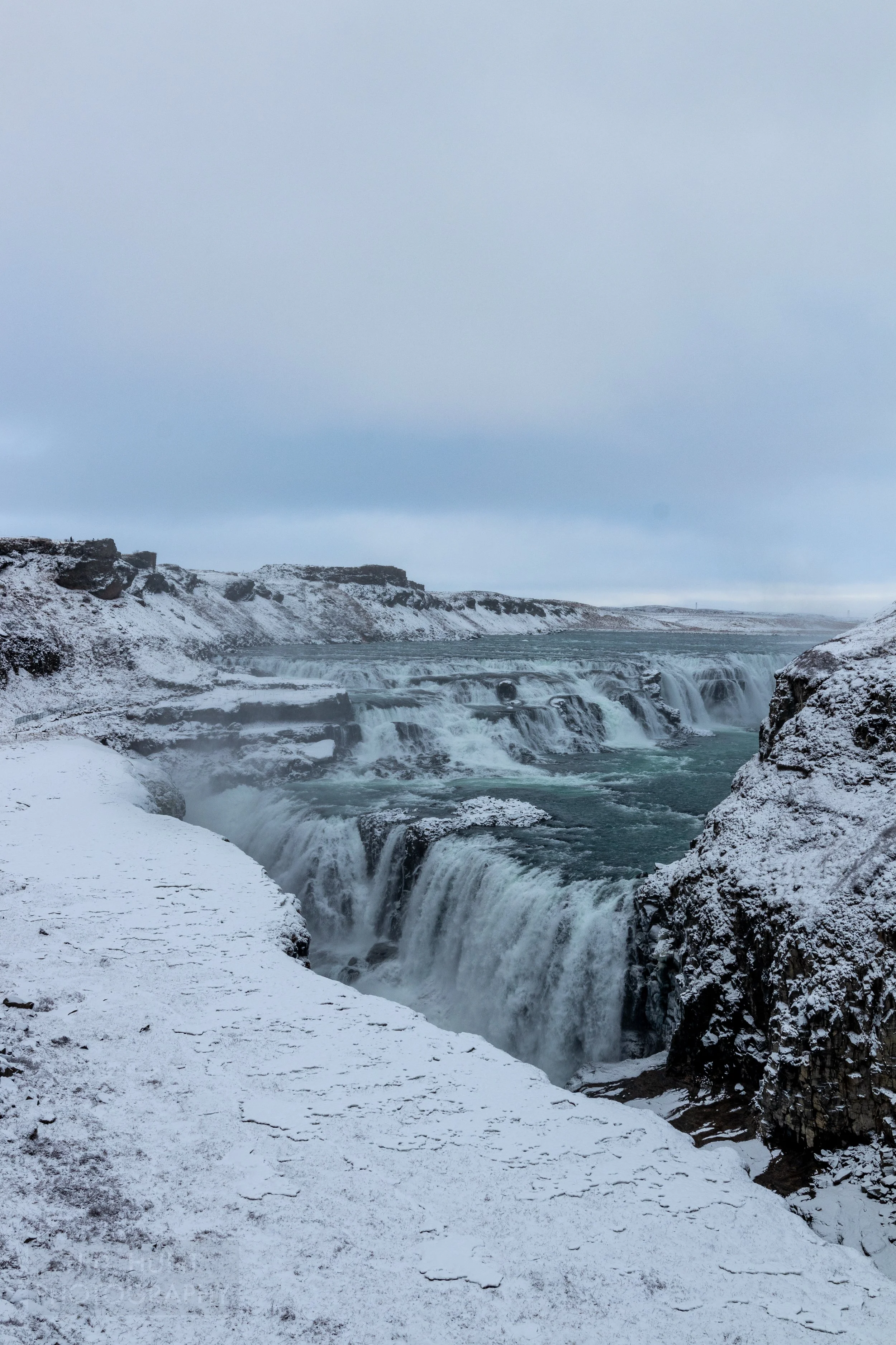 A waterfall, Gullfoss, features dark blue water passing over several tiers of falls surrounded by snow-covered rock cliffs, Iceland.
