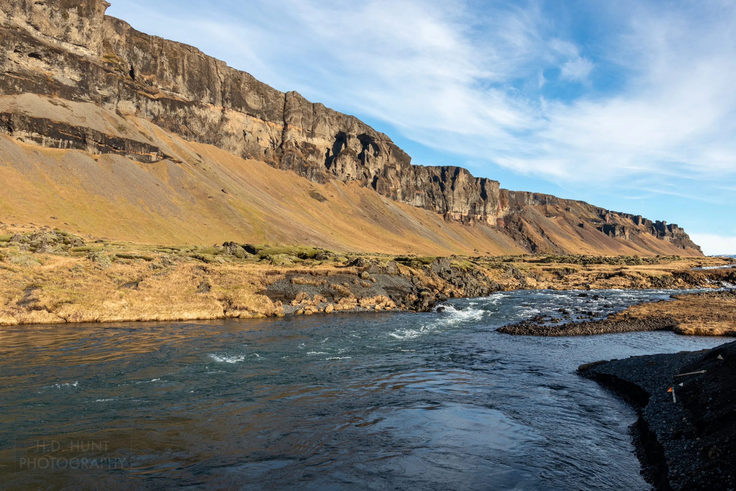 A river current runs beneath tall rocky cliffs, Fossalar, Iceland.