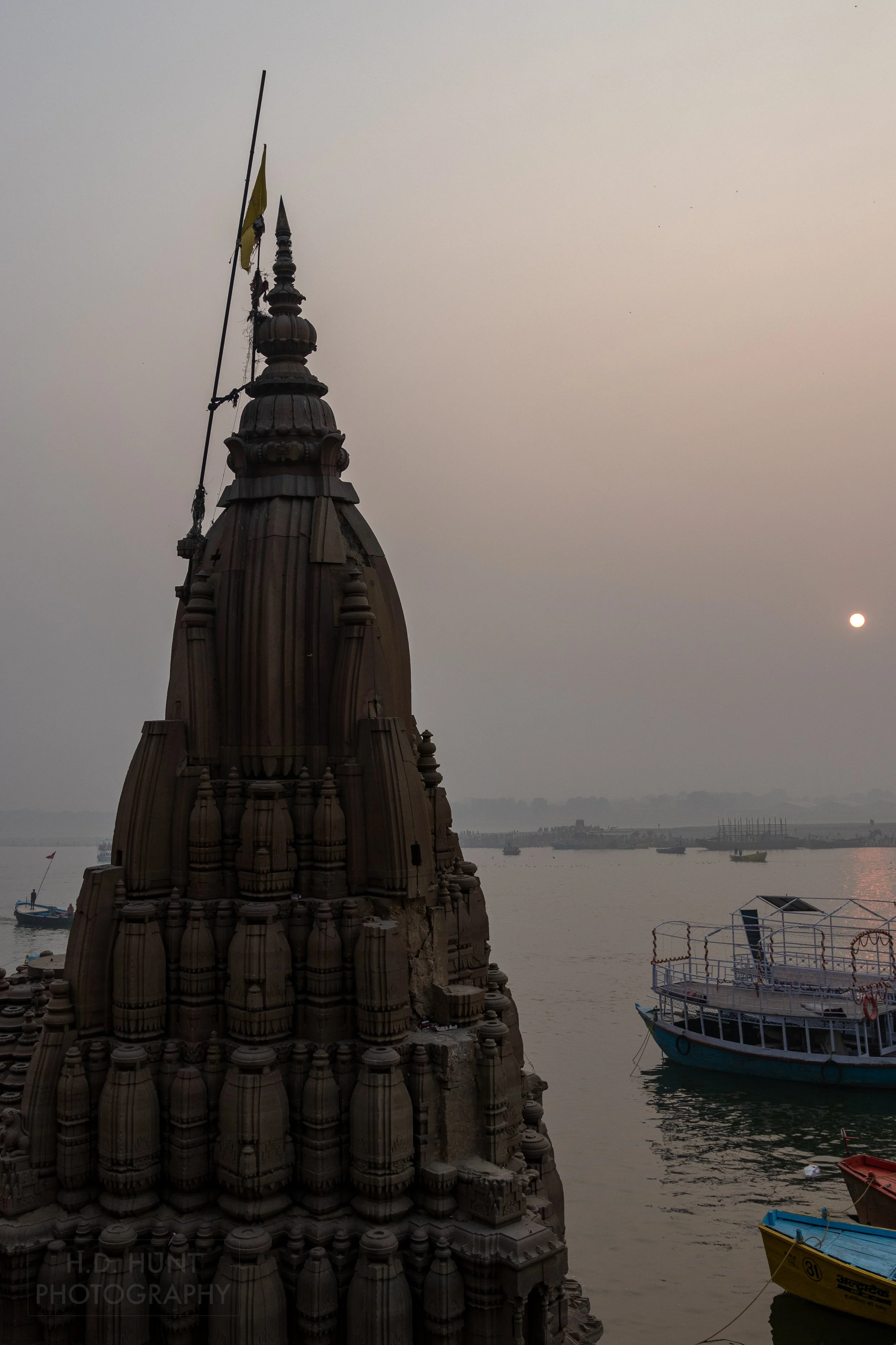 The sun is seen in the background behind a stone tower and the Ganges River, Varanasi, India.