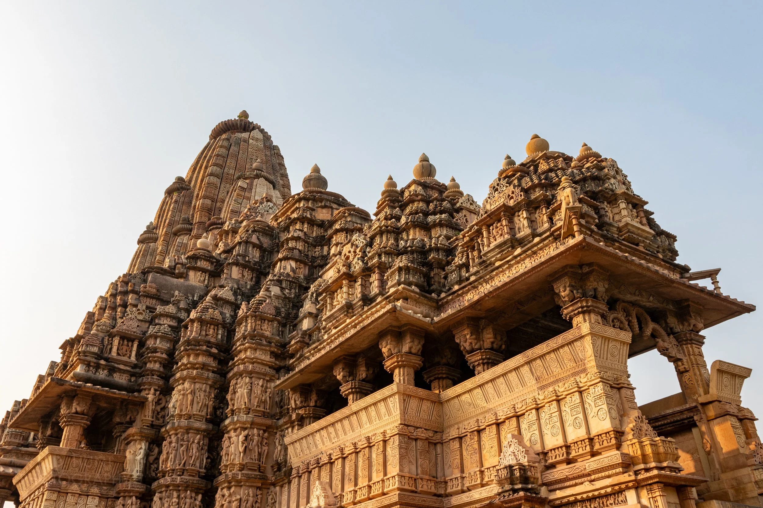 A wide-angle view of the stone carvings adoring Kandariya Mahadeva, Khajuraho Group of Monuments, India, with a tall temple tower rising in the background.
