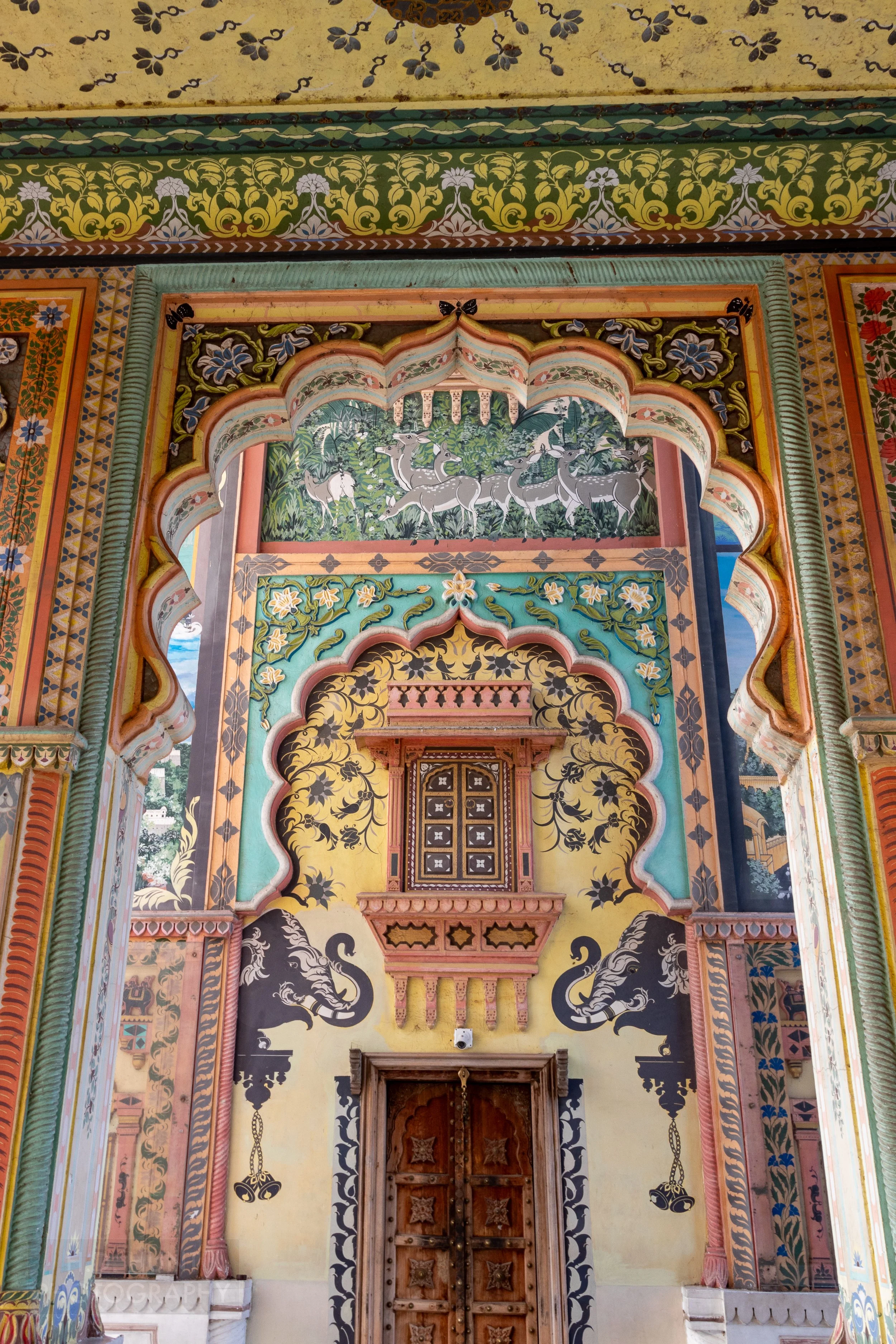 A multi-colored painted wall featuring images of elephants and containing a large wooden door and a small ornamental door, Patrika Gate, Jaipur, India.