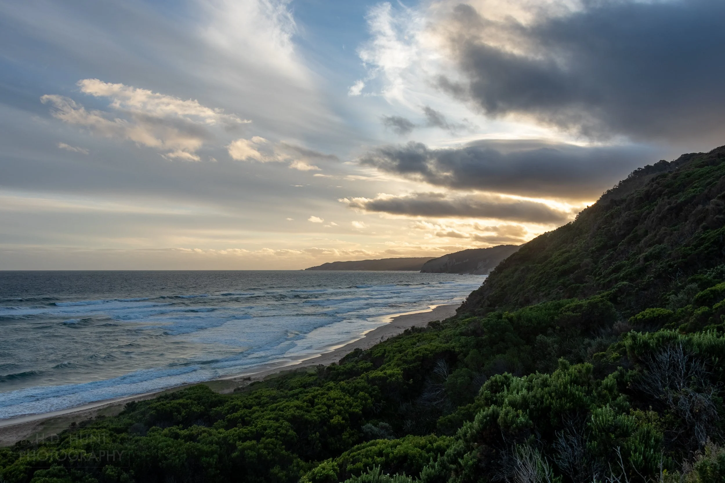 The sun sets behind tall green shrub-covered cliffs and thick dark clouds near Johanna, Victoria, Australia.