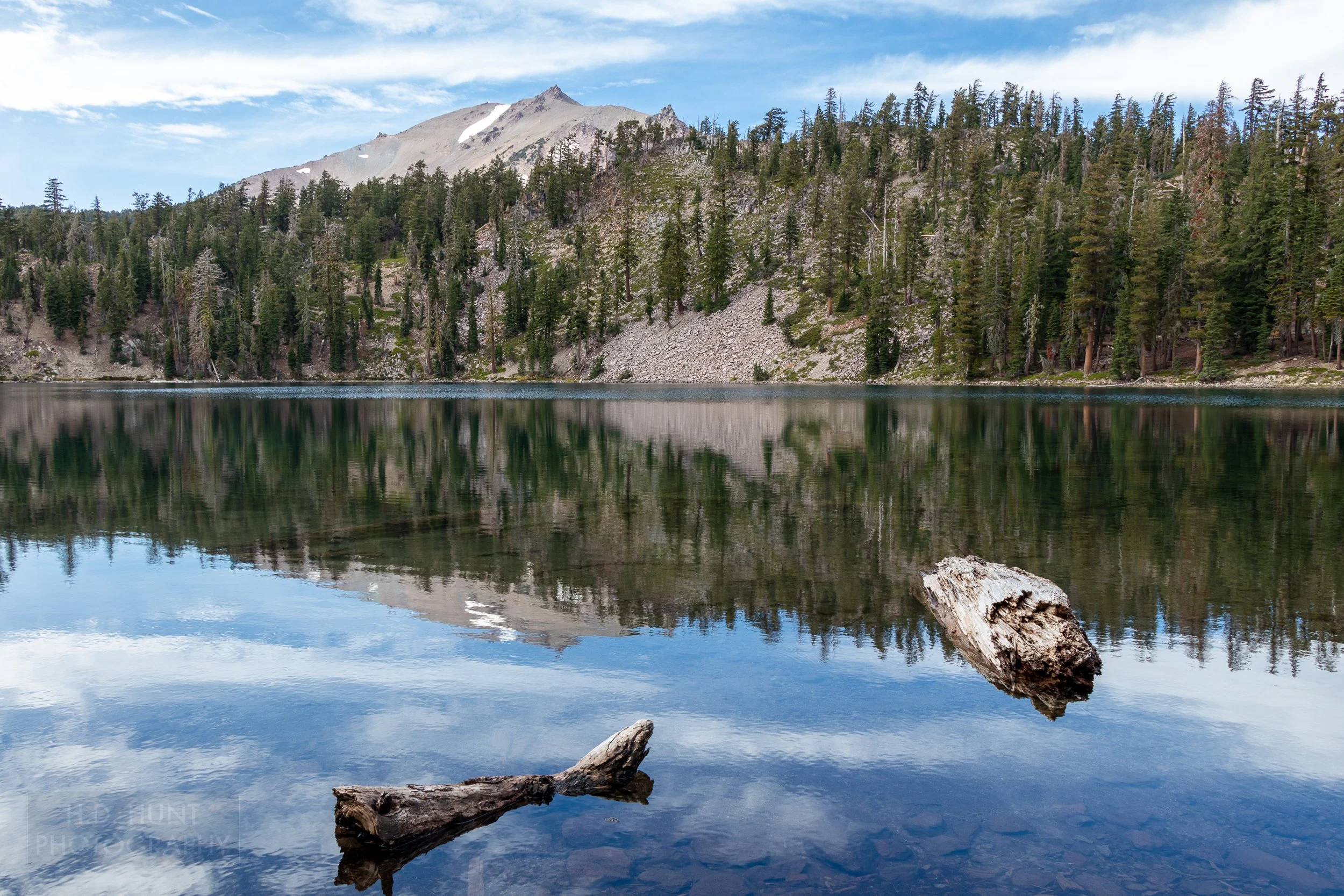 Tree trunk pieces protrude from a clear blue lake reflecting a distant mountain in Lassen Volcanic National Park, California, United States.