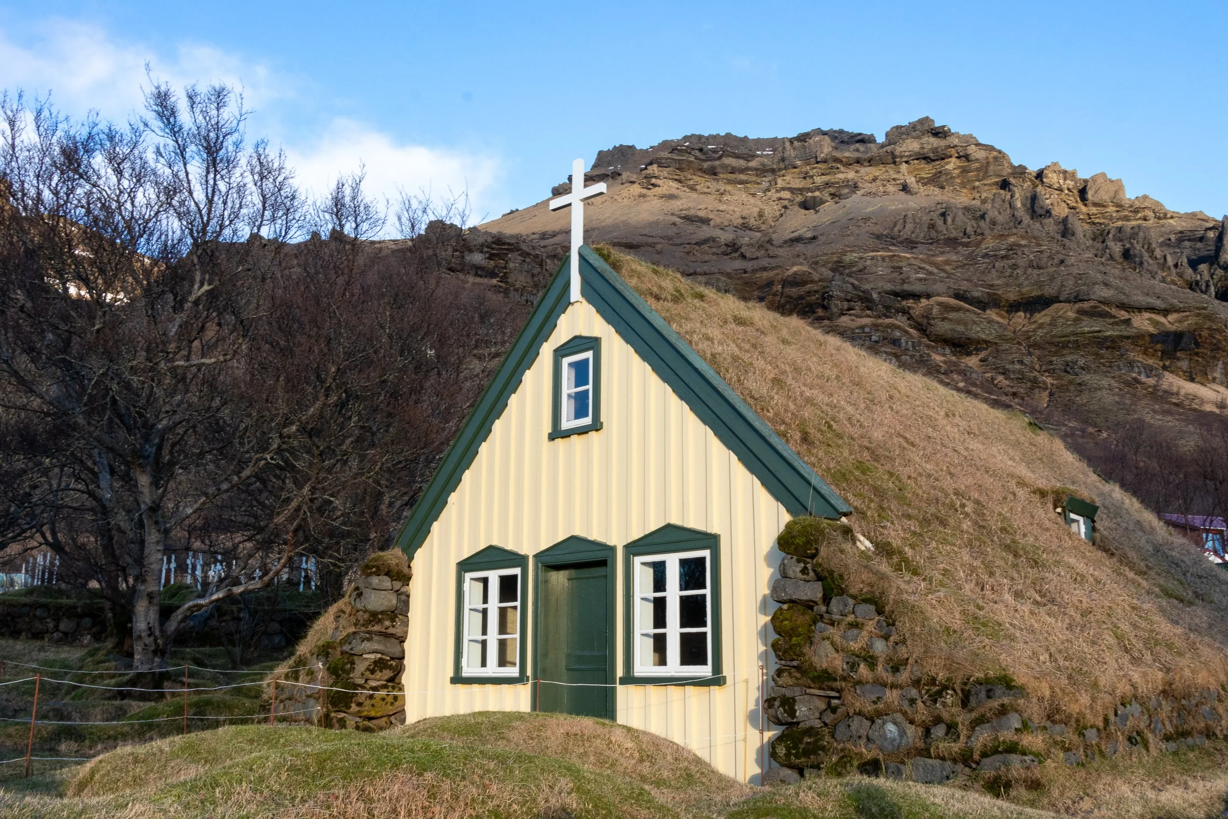 A yellow and green church building with a turf grass roof sits in front of a large rocky hill, Hof, Iceland.